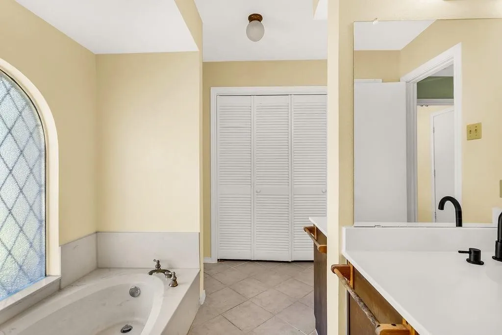 Soaking tub with a decorative arched window, dual basin vanity with dark bronze fixtures, and tile flooring