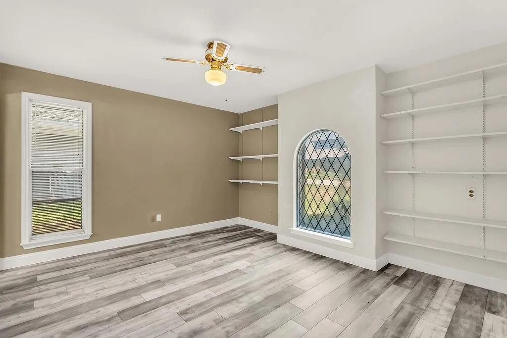 Wood-finish flooring throughout, featuring a gold-tone ceiling fan with integrated lighting, a rectangular window, and a prominent arched window with decorative muntins