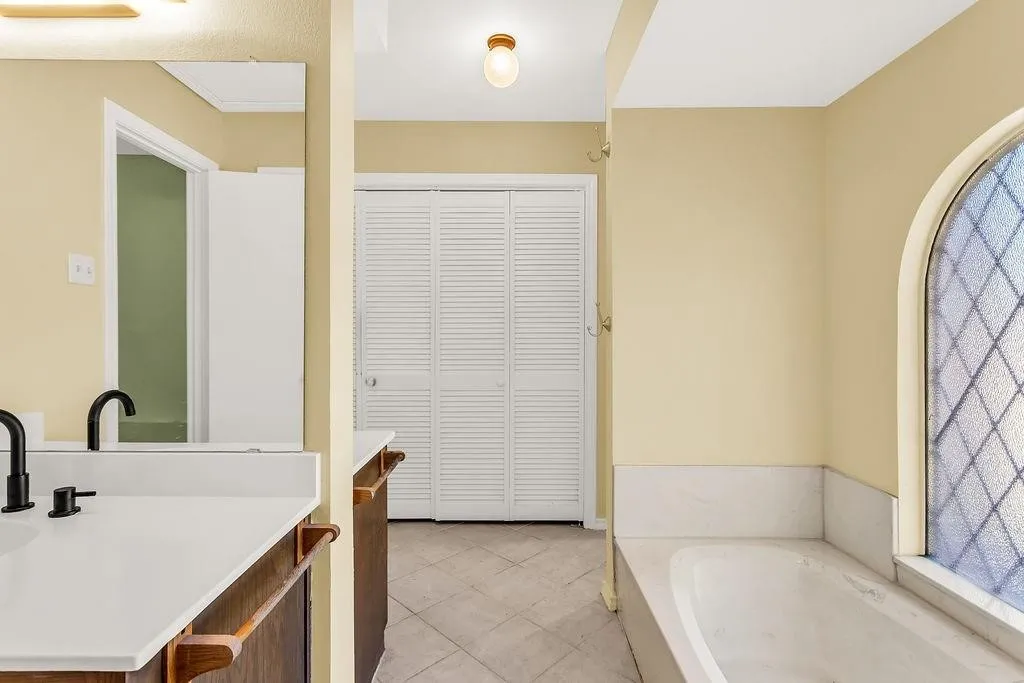 Spa-inspired primary bathroom featuring a soaking tub with an arched window, dual vanity with wood-finish cabinetry and black hardware, tile flooring, and direct access to an additional room through a closet door