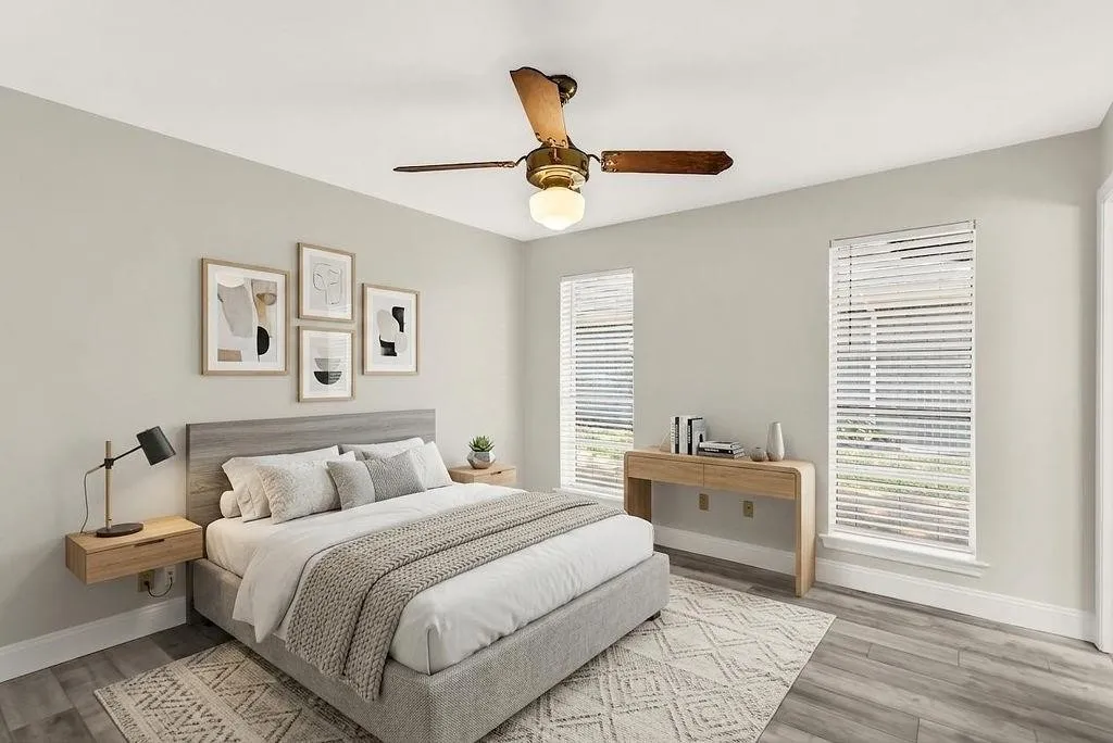 Wood-finish flooring with light-toned walls, two windows with blinds, a ceiling fan with integrated lighting, and a built-in wood-look headboard