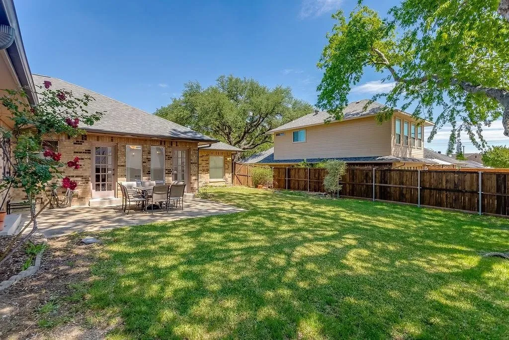 Expansive backyard oasis featuring a manicured lawn, mature shade trees, and a covered patio with dining area