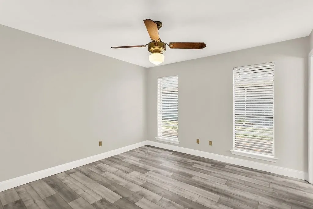Light gray wall paint, wood-finish flooring, two large windows, white window blinds, and a ceiling fan with light fixture