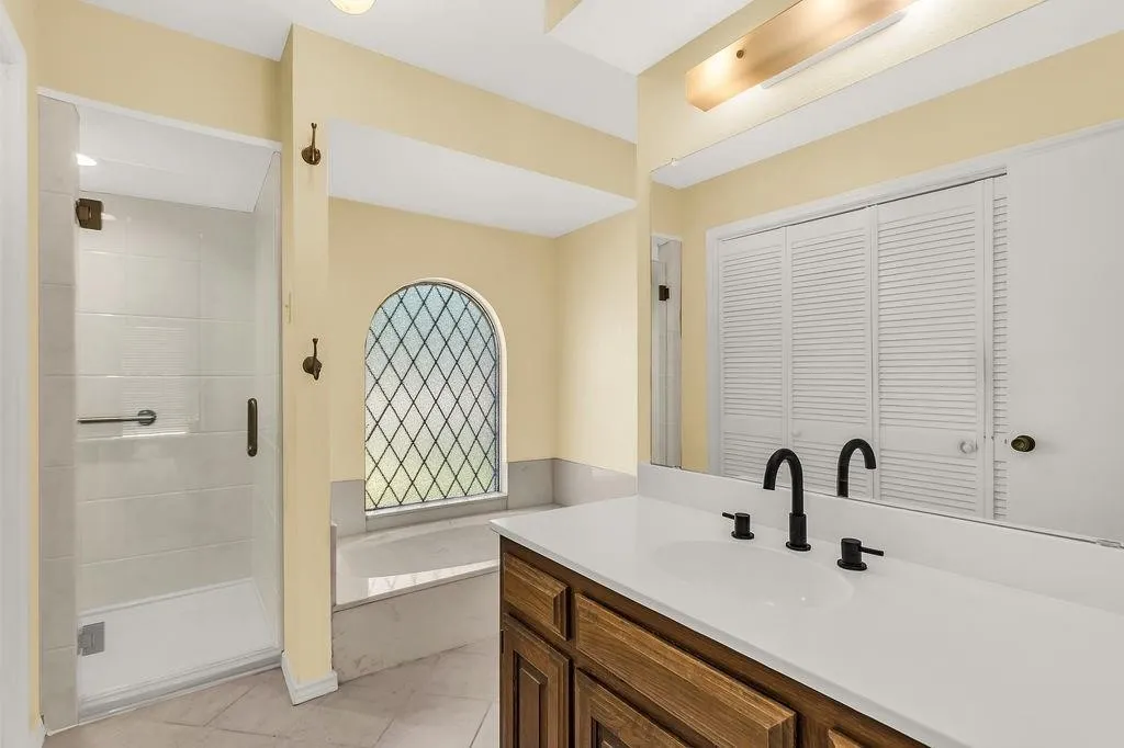 Glass-enclosed shower with white tile surround, built-in bathtub with arched window, wood vanity with white countertop, dual sinks with matte black fixtures, and louvered closet doors