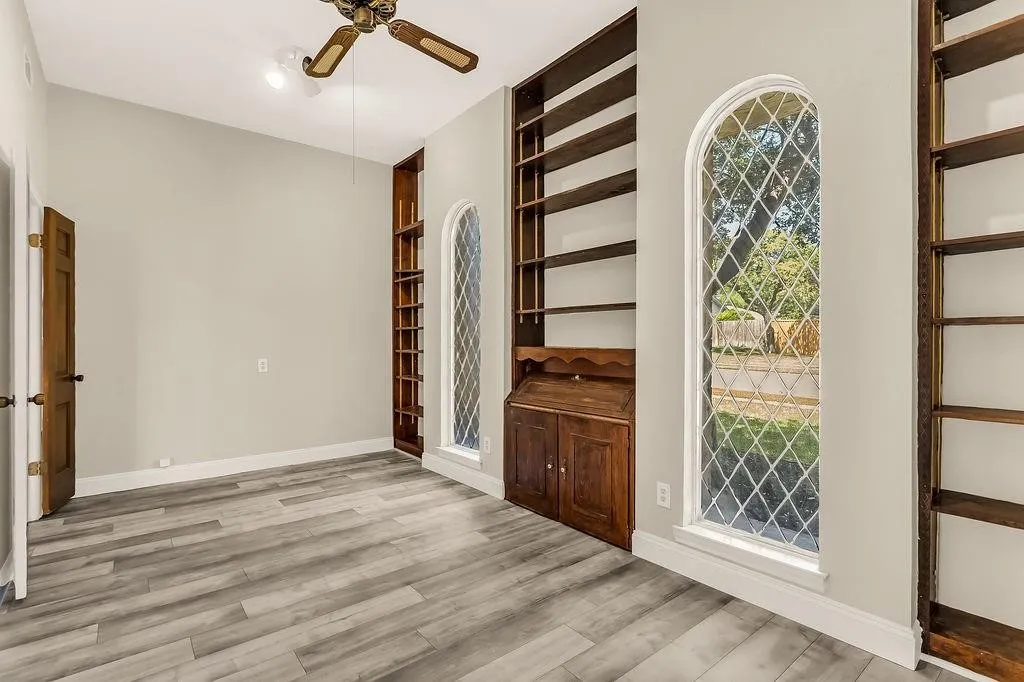 Wood-finish flooring, two arched leaded-glass windows, built-in shelving, built-in desk, and ceiling fan with integrated lighting