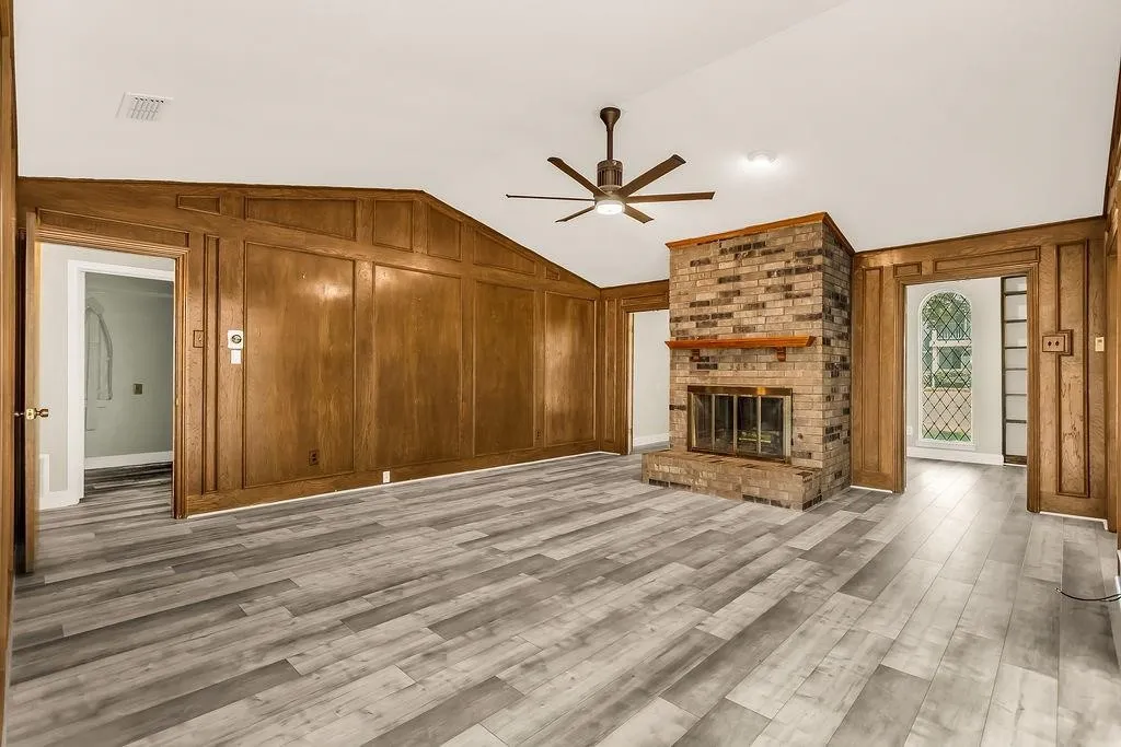 Vaulted ceiling, wood-finish flooring, paneled walls, brick fireplace with wood mantel, and a ceiling fan