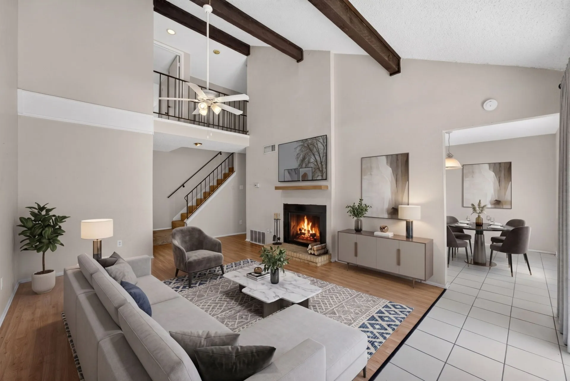 Vaulted ceiling with exposed wood beams, wood-finish flooring, fireplace with brick surround and wood mantel, upper-level balcony with metal railing, and a staircase with wood treads and metal railing
