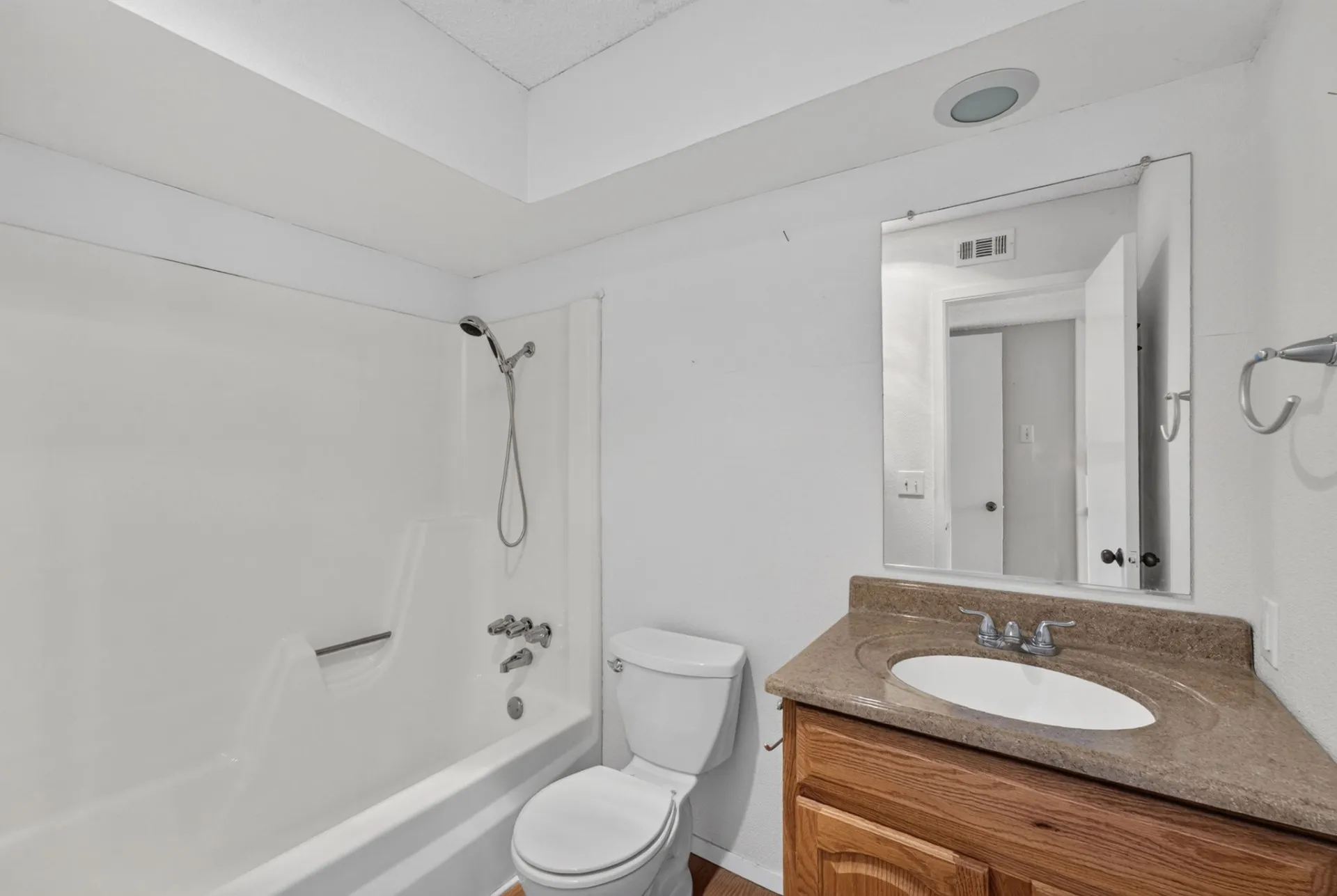White bathtub and shower combination with a showerhead, a toilet, a wood-finish vanity with an integrated sink, and a wall-mounted mirror