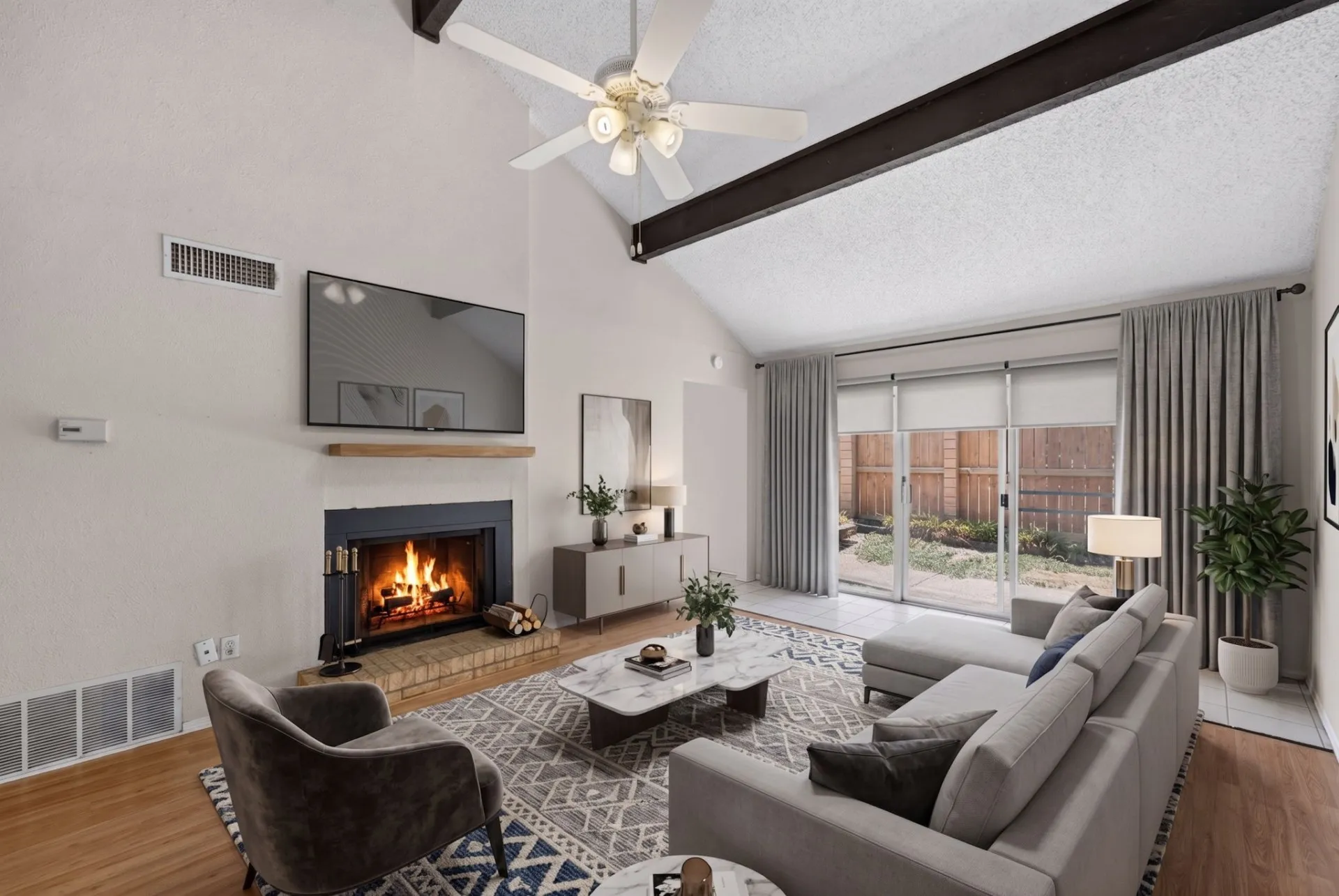 Vaulted ceiling with exposed beam, brick-surround fireplace with wood mantel, wood-finish flooring, full-light sliding patio door, and ceiling fan with integrated lighting