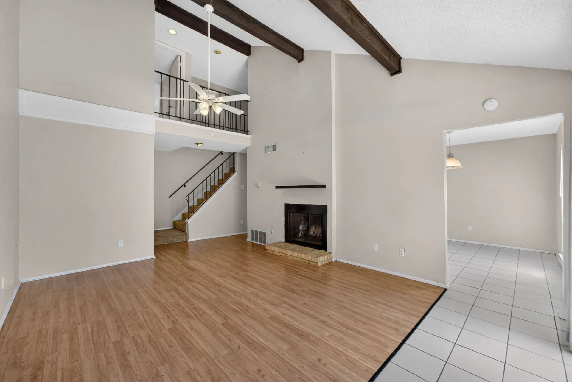 Vaulted ceiling with exposed beams, wood-finish flooring, fireplace with brick hearth, interior balcony with metal railing, and a ceiling fan