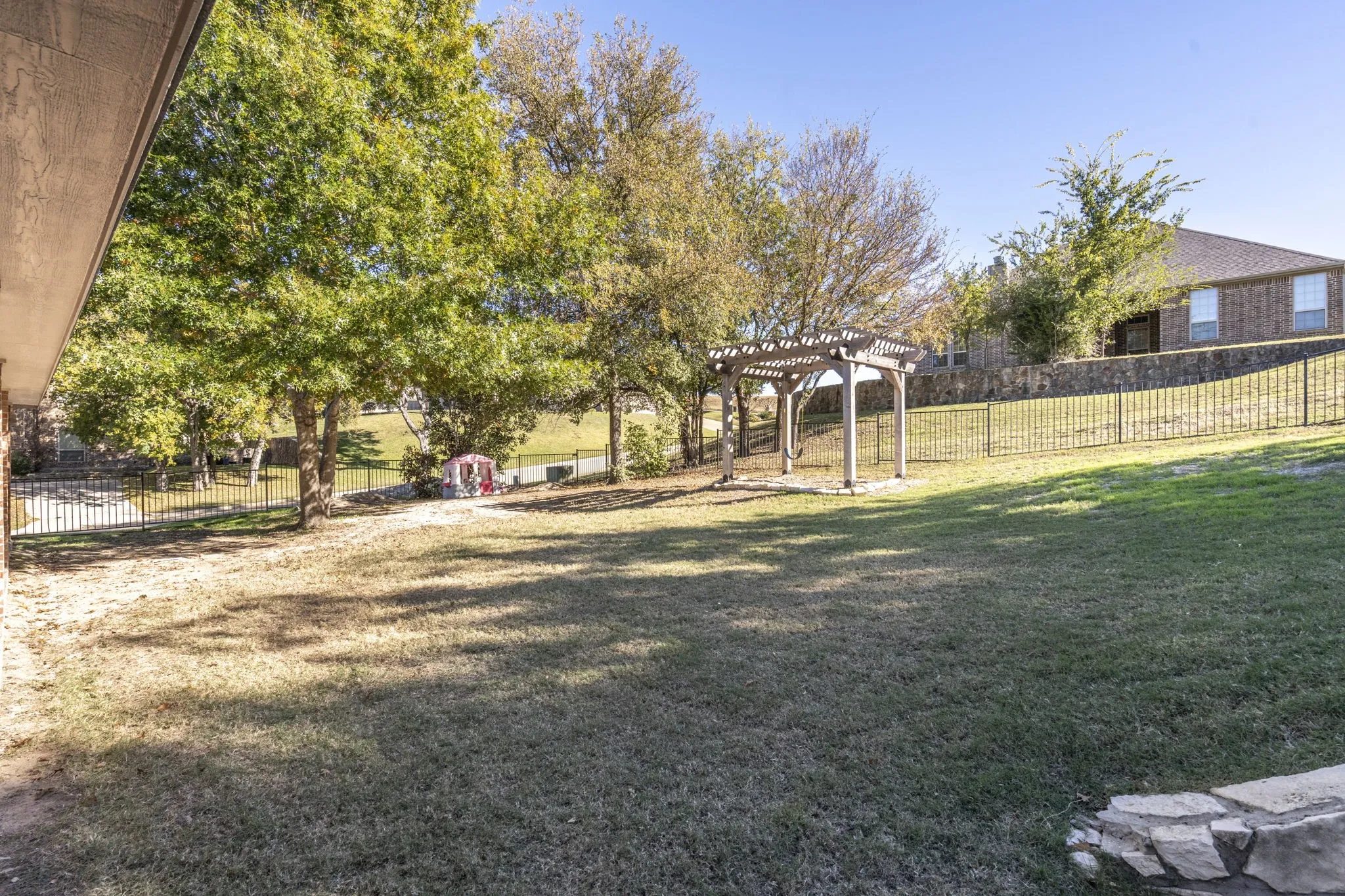 Spacious backyard featuring a pergola, mature trees, and a black wrought iron fence