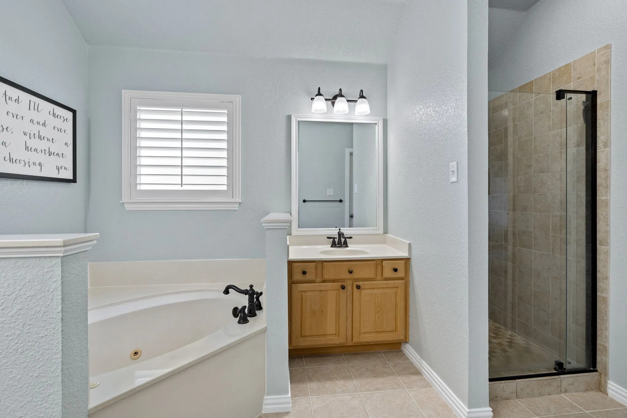 Corner soaking tub, wood vanity with a light-colored countertop, framed mirror, three-light vanity fixture, and a tiled walk-in shower with a glass enclosure