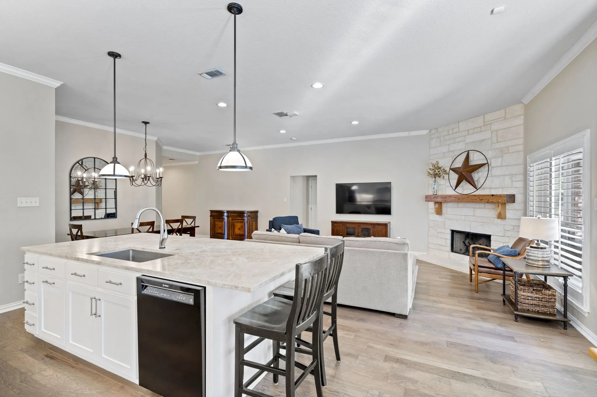 Spacious kitchen island with a built-in sink and dishwasher, pendant lighting, stone-clad fireplace with a wood mantel, and wood-finish flooring