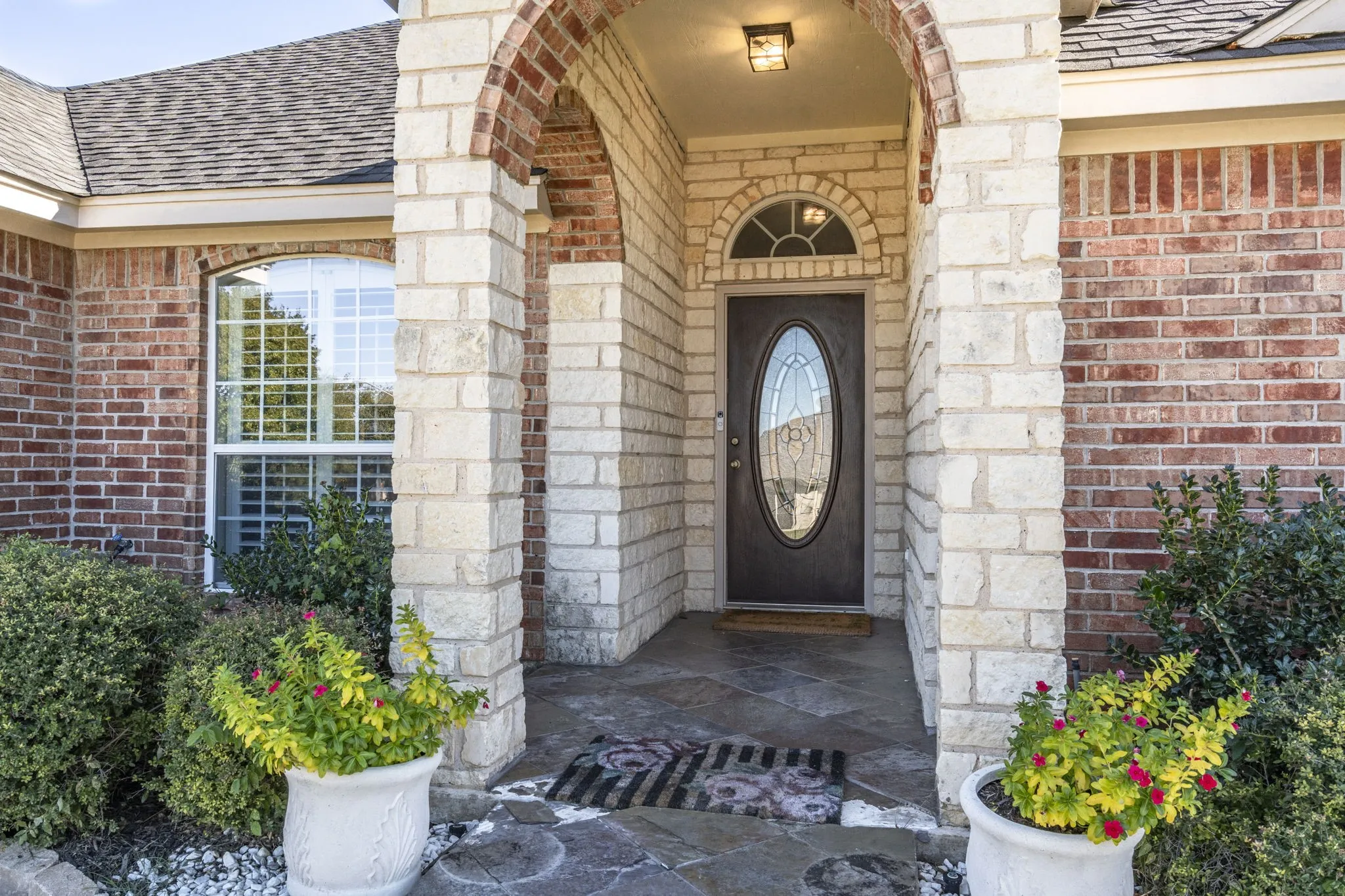 Arched entryway featuring brick and stone masonry, a dark paneled door with an oval glass insert, a transom window, and a stone-tiled porch