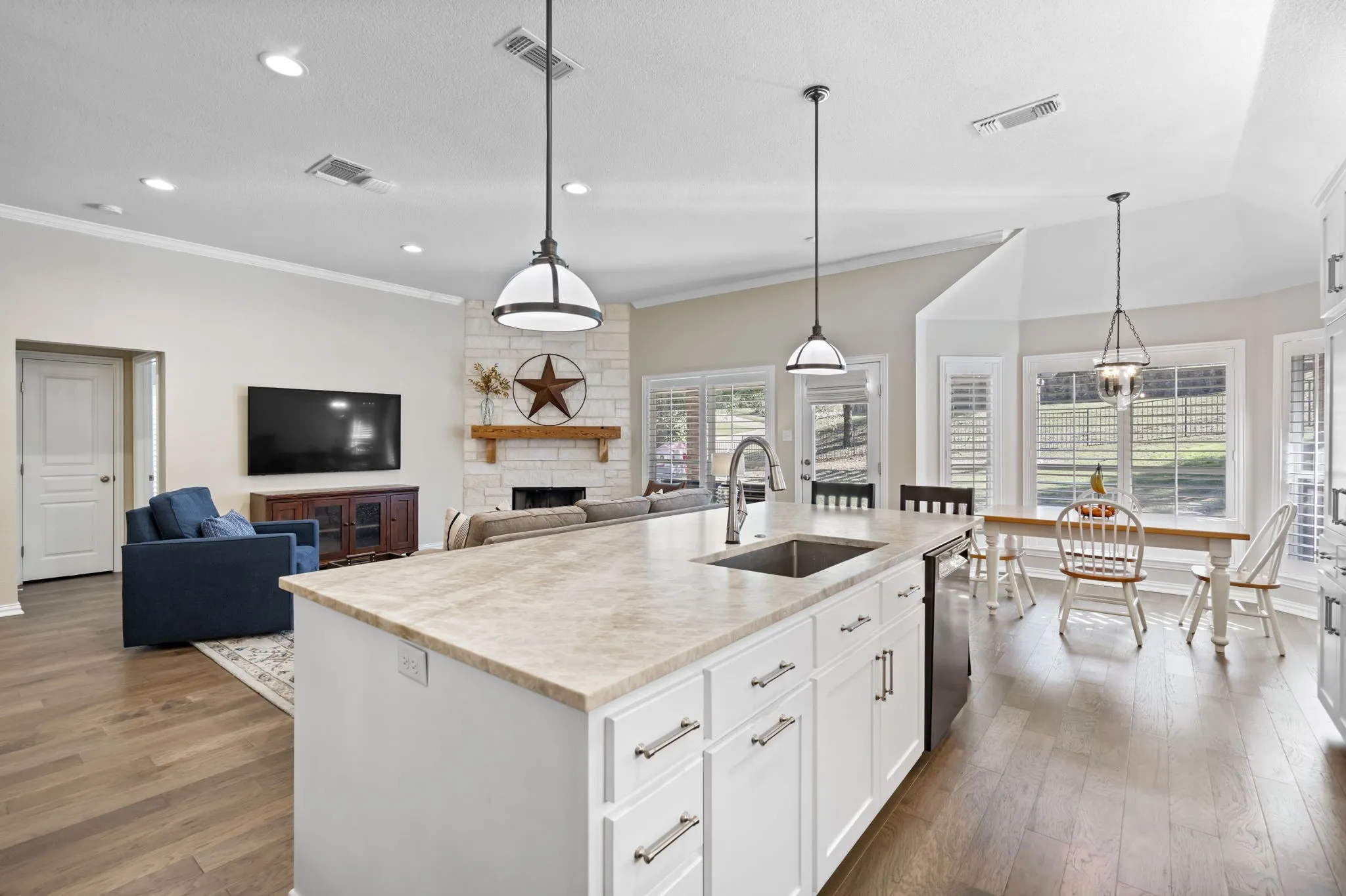 Kitchen island with an undermount sink, pendant lighting, wood-finish flooring, white cabinetry, and a stone fireplace