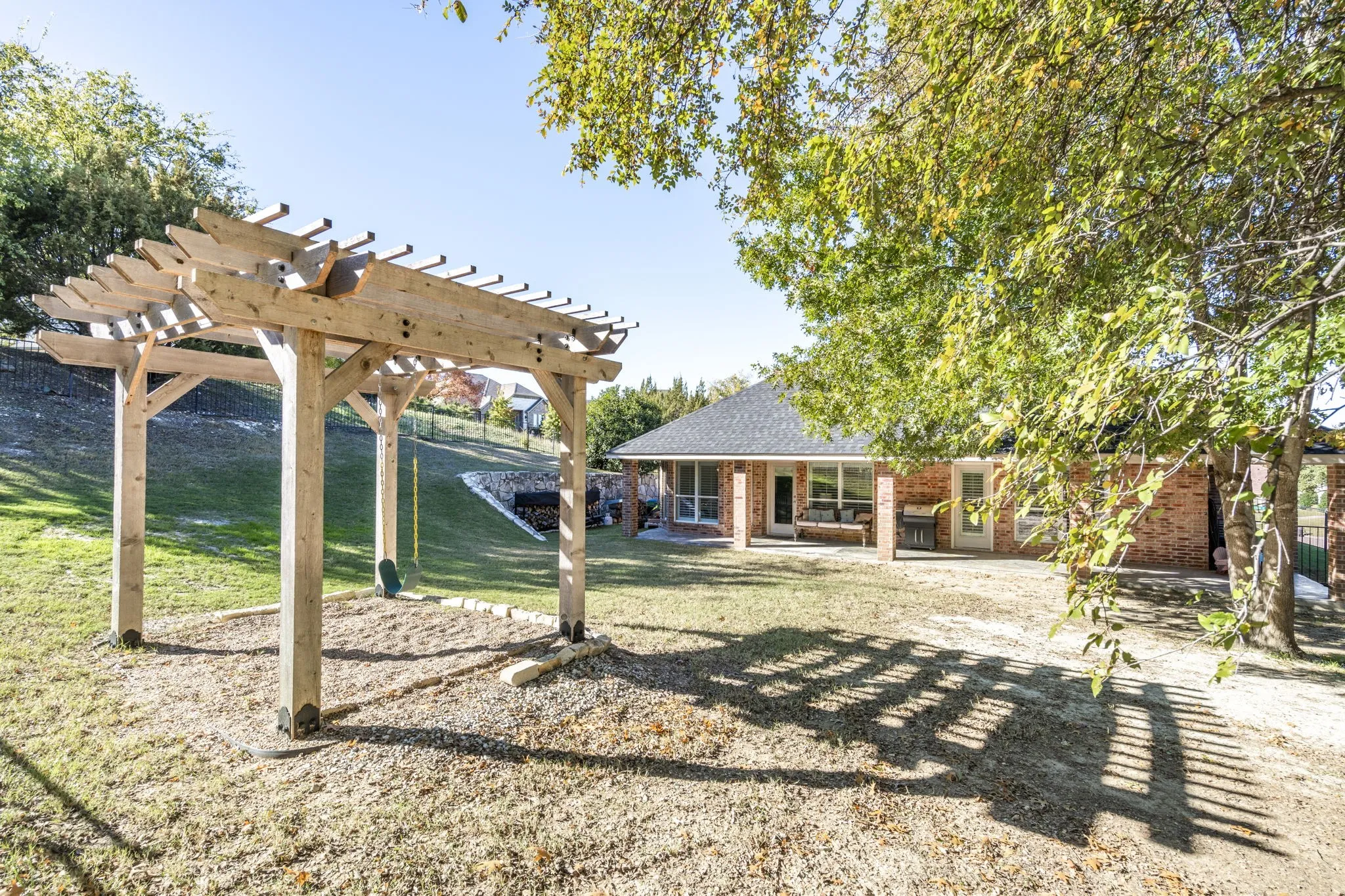 Wood pergola over a gravel base, expansive lawn, mature foliage, brick exterior, and covered patio