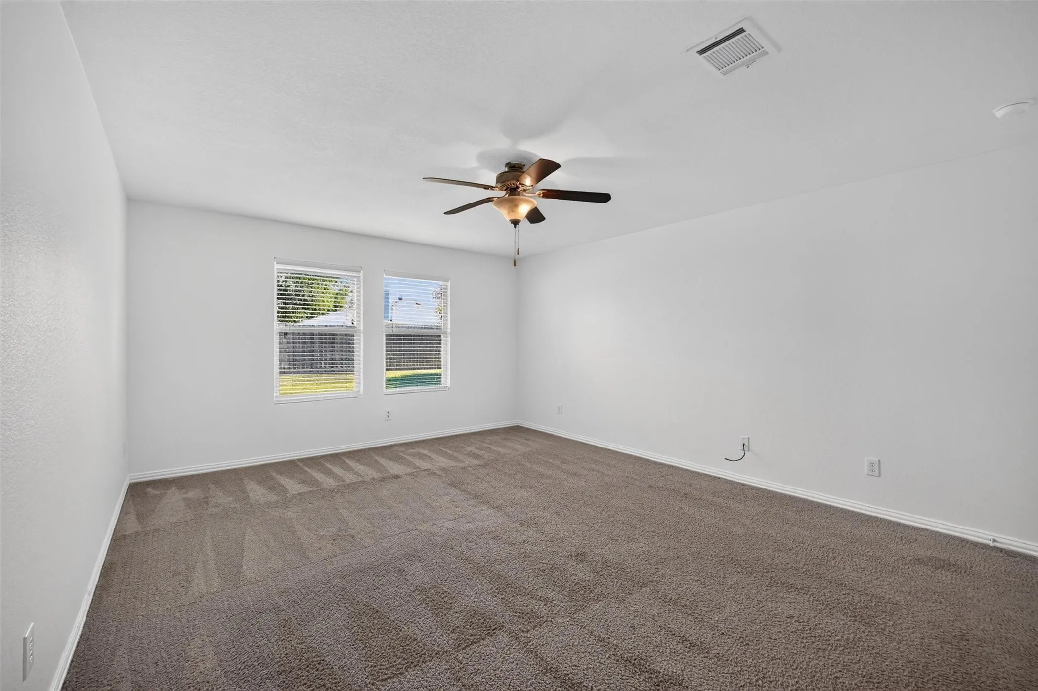 Spacious room featuring neutral carpeting, white painted walls, two windows with blinds, and a ceiling fan