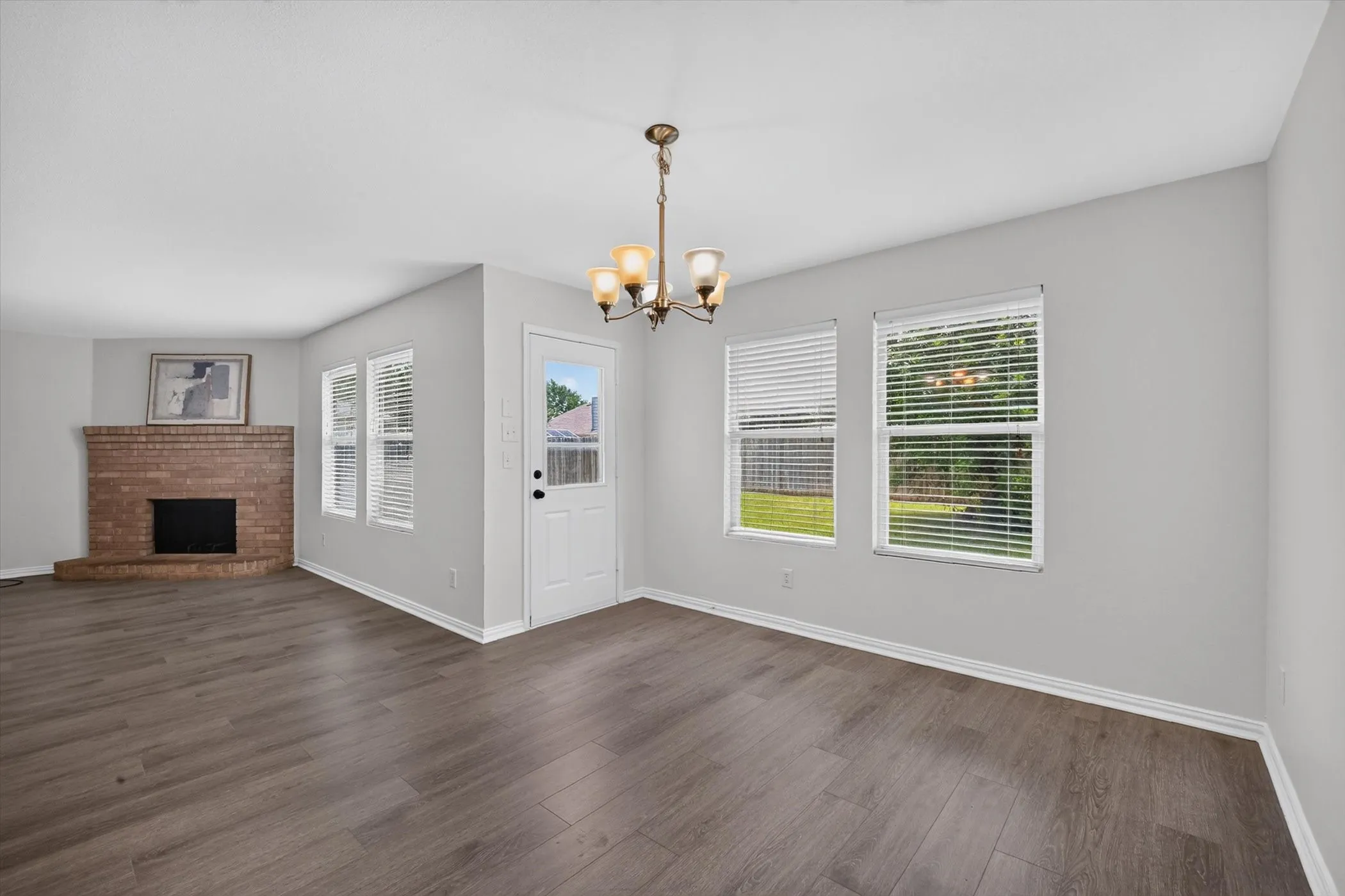 Wood-finish flooring, brick fireplace, multiple windows, white paneled door, and a bronze chandelier with glass shades
