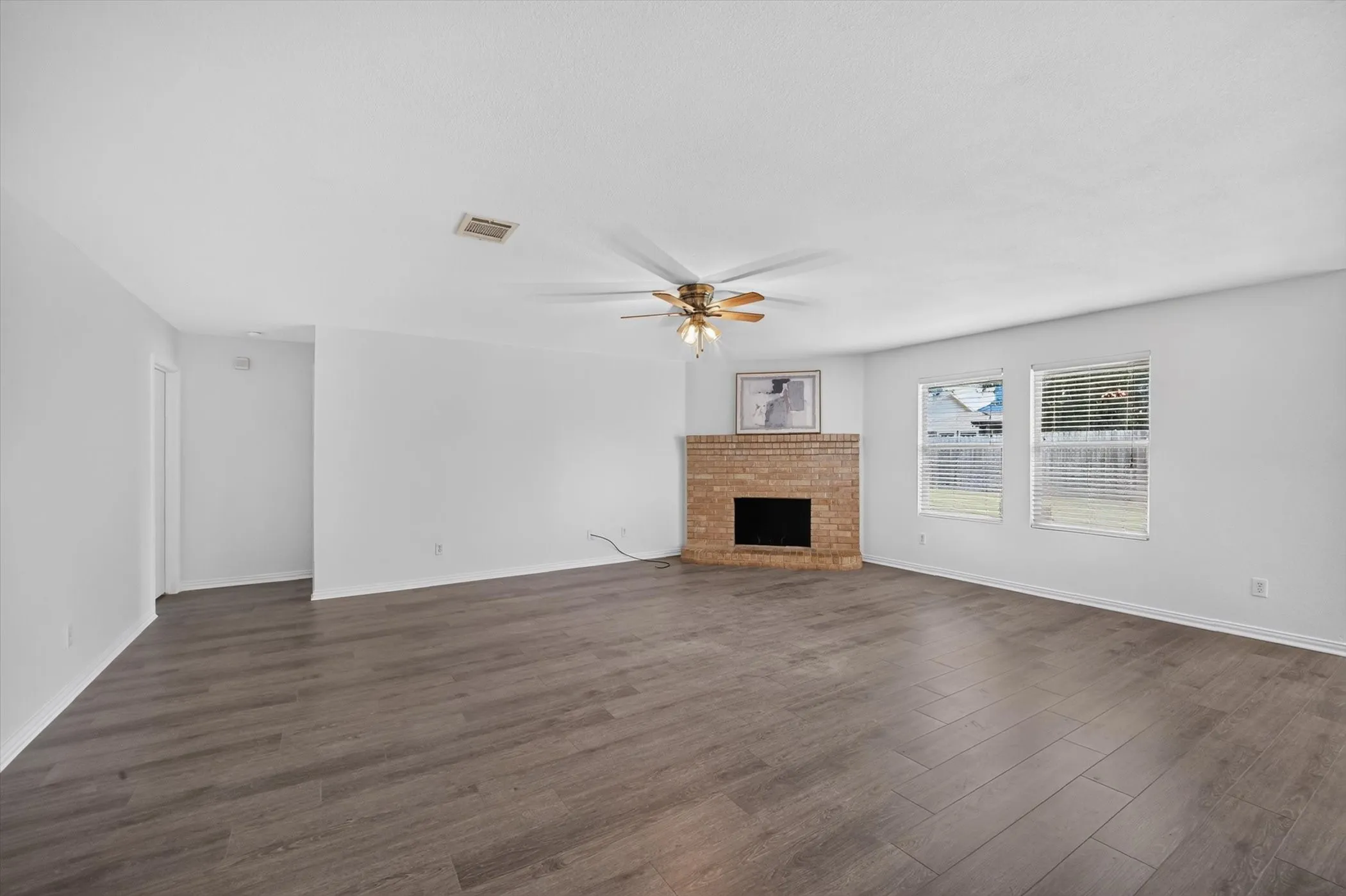 Spacious living area featuring wood-finish flooring, a corner brick fireplace, and two​​‌​​​​‌​​‌‌​‌‌​​​‌‌​‌​‌​‌​​​‌​​ windows