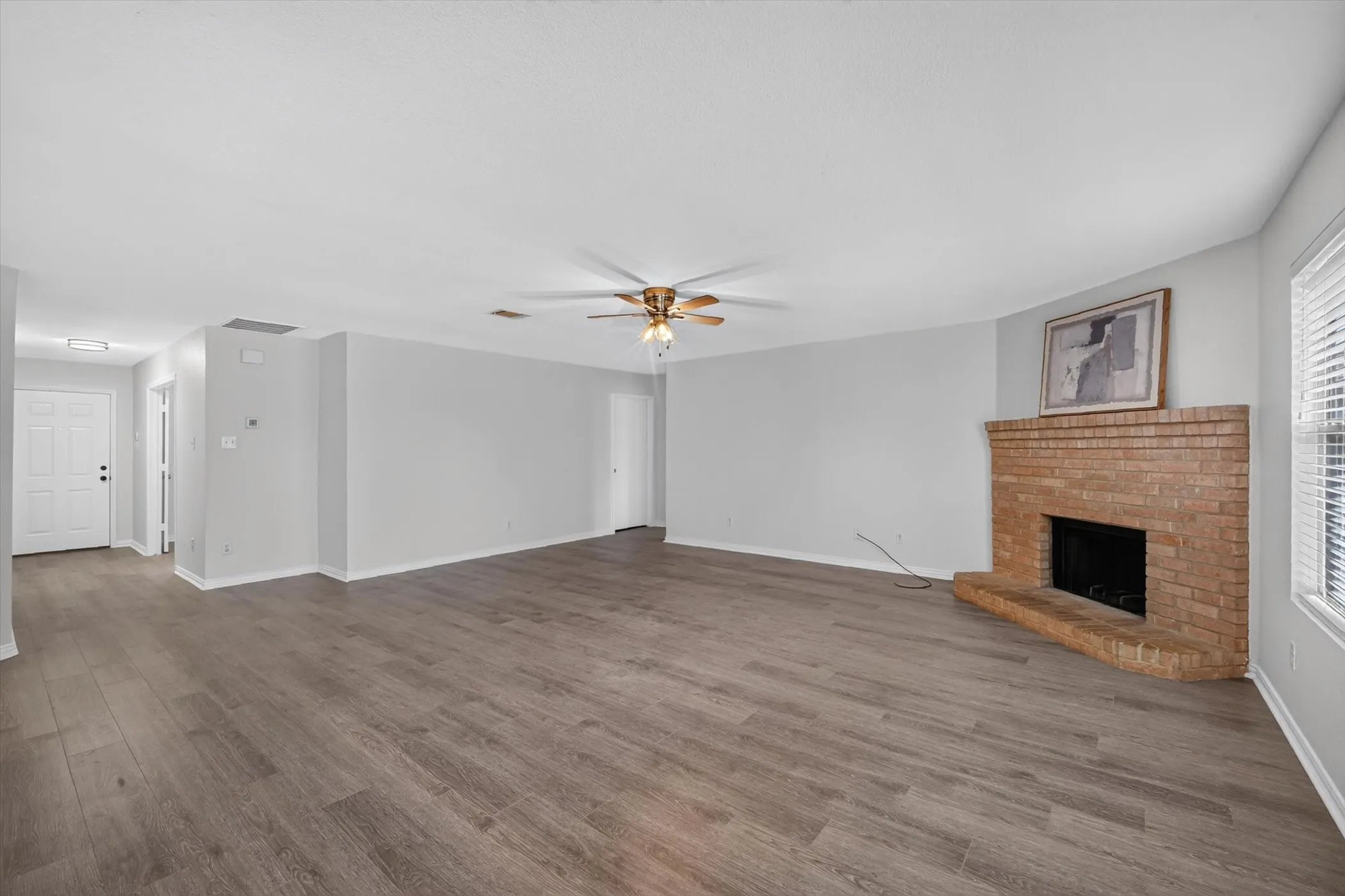 Spacious interior with wood-finish flooring, a brick fireplace, ceiling fan, and window with blinds