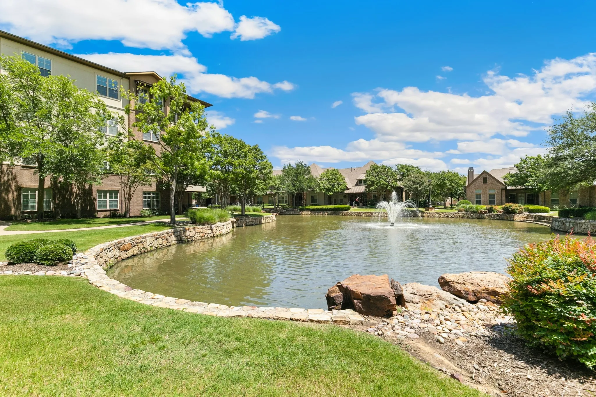 Lush green lawn with a retaining wall, mature trees, and a central pond featuring a fountain