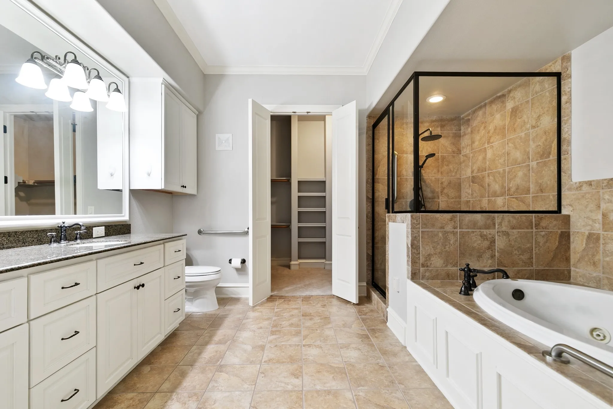 Dual vanity with undermount sinks and dark countertop, framed mirror with overhead lighting, tiled walk-in shower with black-framed glass enclosure, jetted soaking tub, and tile flooring