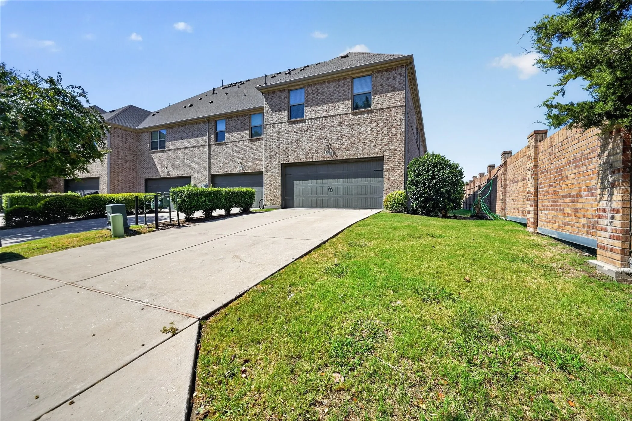 Brick exterior, dark shingle roof, attached garage with dark door, concrete driveway, and manicured landscaping