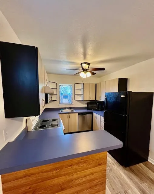 U-shaped kitchen featuring wood-finish flooring, two-tone cabinetry, dark countertops, and a ceiling fan with integrated lighting