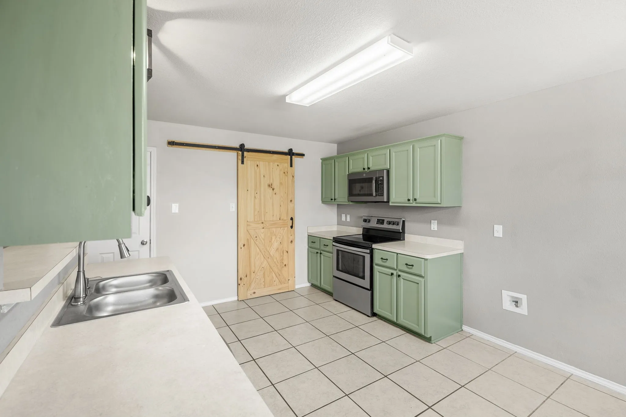 Kitchen featuring light green cabinetry, stainless steel appliances, a double basin sink, light-toned countertops, and a wood sliding barn door