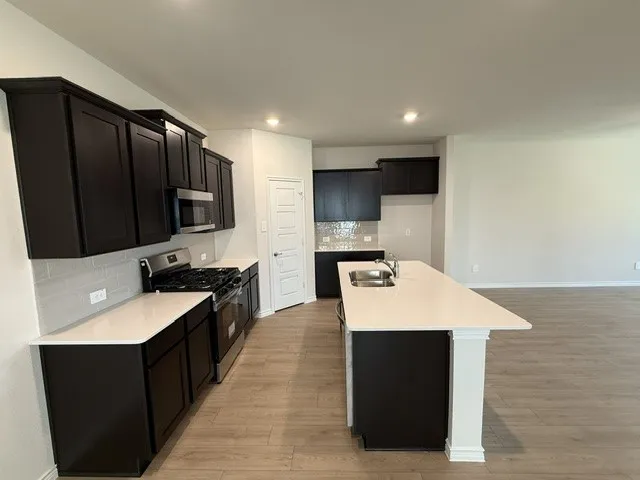 Kitchen featuring dark wood-finish cabinetry, light countertops, stainless steel appliances, a tiled backsplash, and an island with an integrated sink
