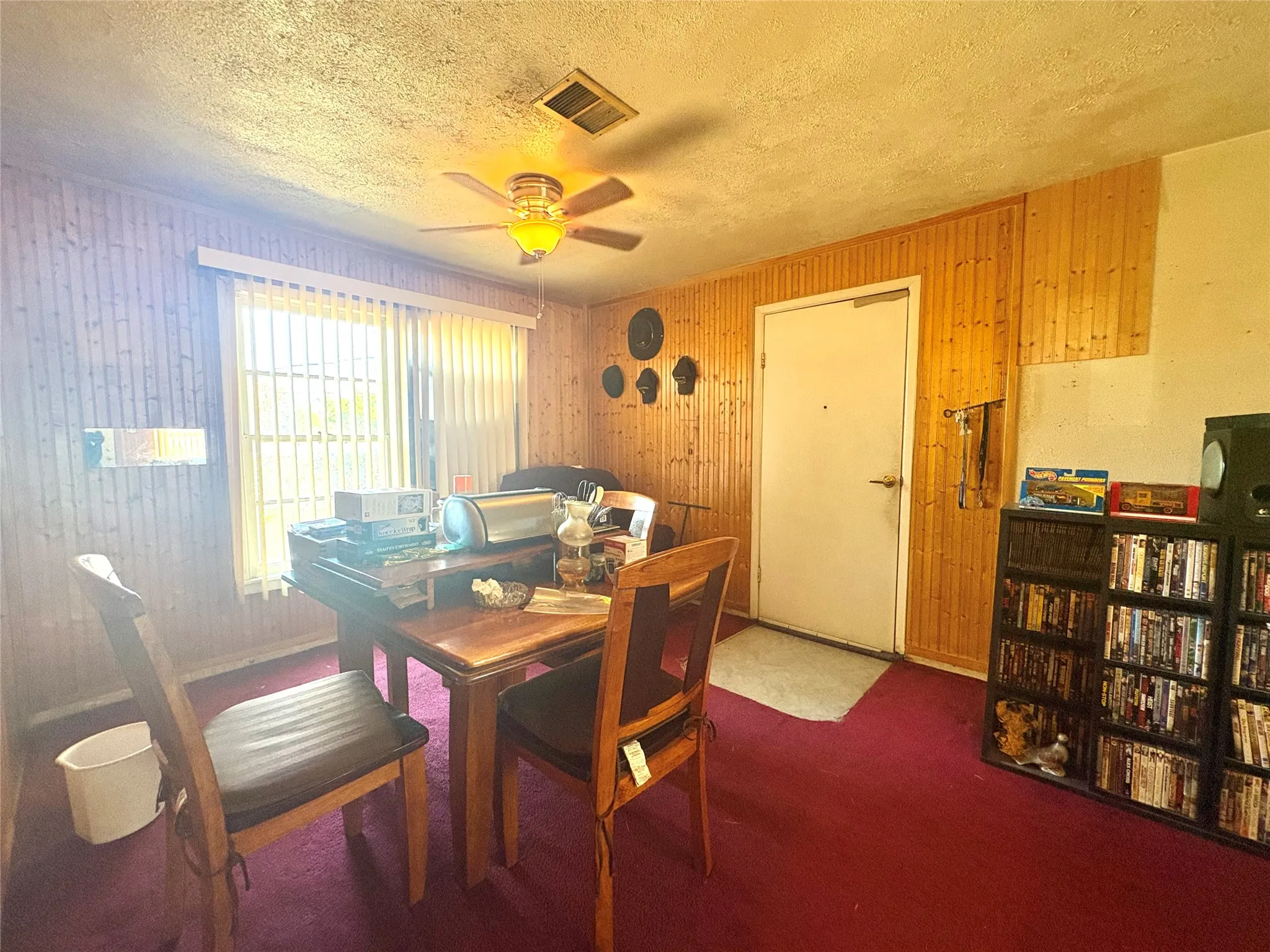 Wood paneling on walls, ceiling fan with integrated lighting, large window with blinds, interior door, and red carpeting