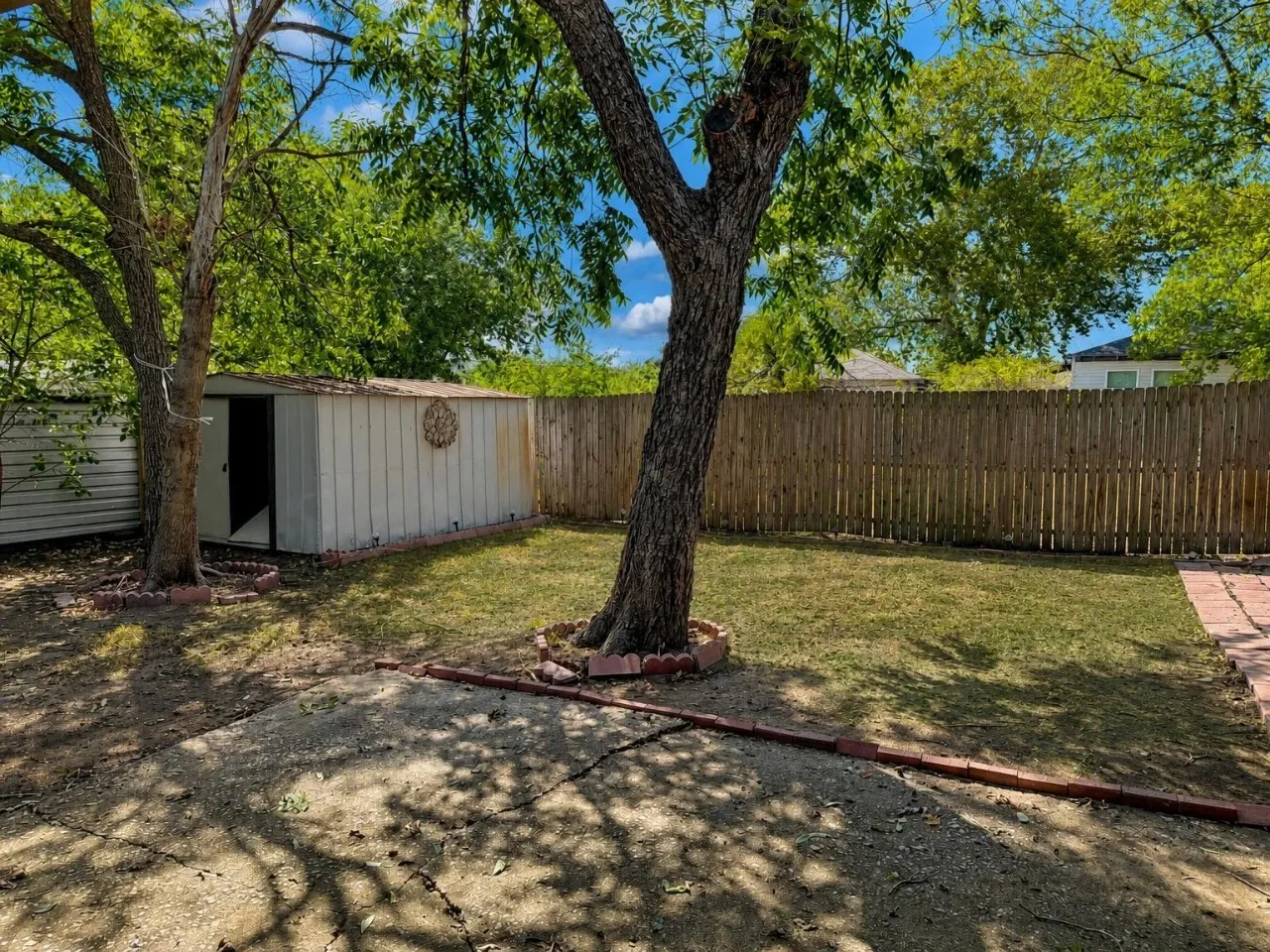 Fenced backyard with mature trees, a storage shed, and a brick-lined concrete patio