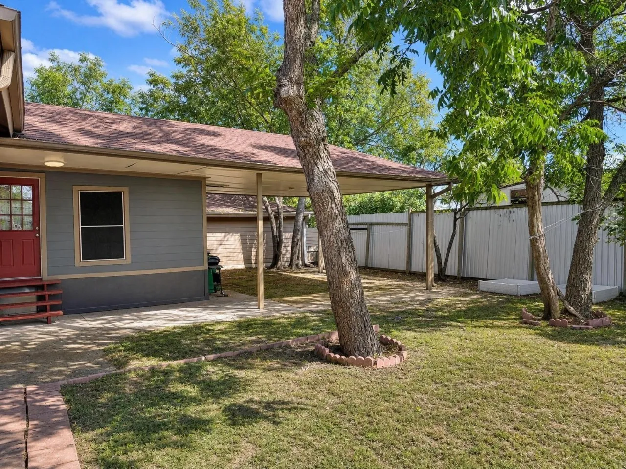 Covered patio, red entry door, shingled roof, mature trees, metal fencing