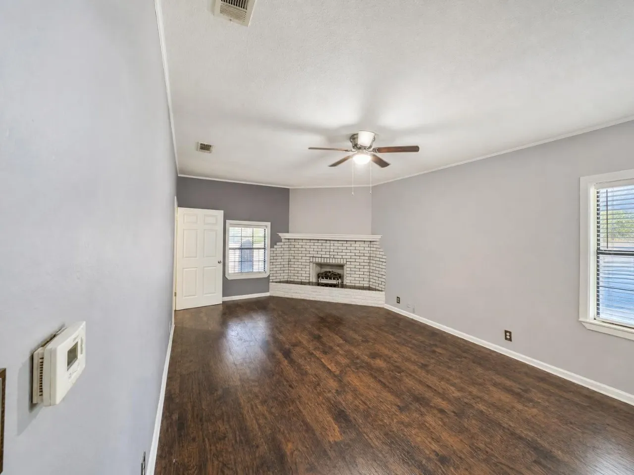 Dark wood-finish flooring, white brick fireplace, ceiling fan, white paneled door, and window with blinds