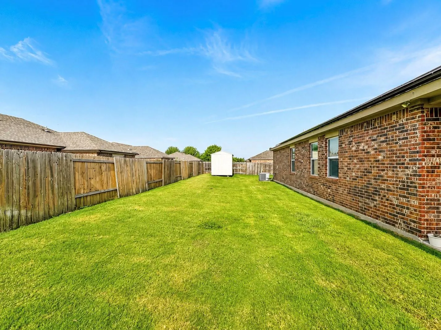 Expansive backyard featuring a manicured lawn, wood privacy fencing, a white storage shed, and a brick exterior