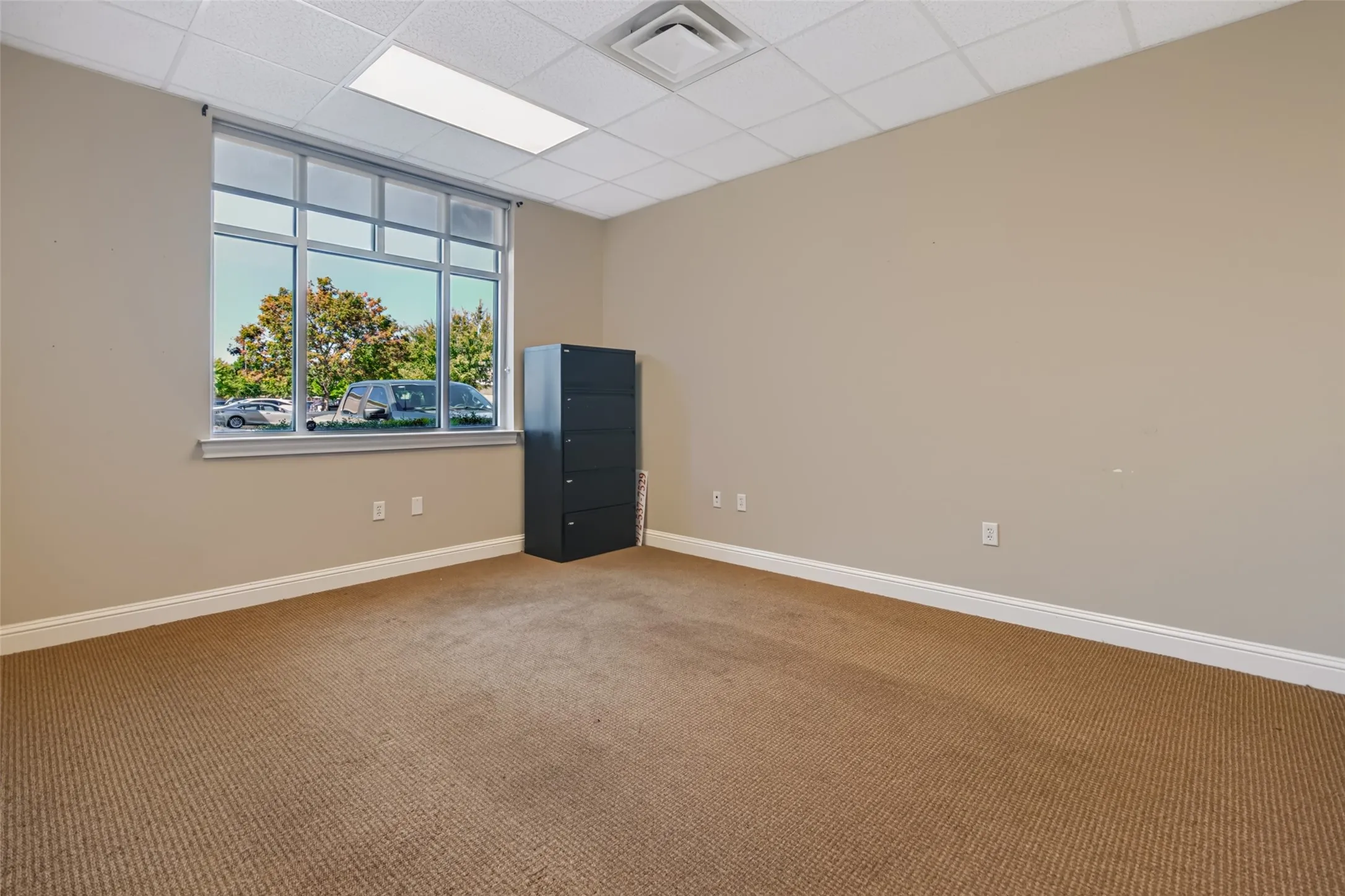 Carpeted room featuring a large window with multiple panes, drop ceiling with integrated lighting, and neutral wall tones