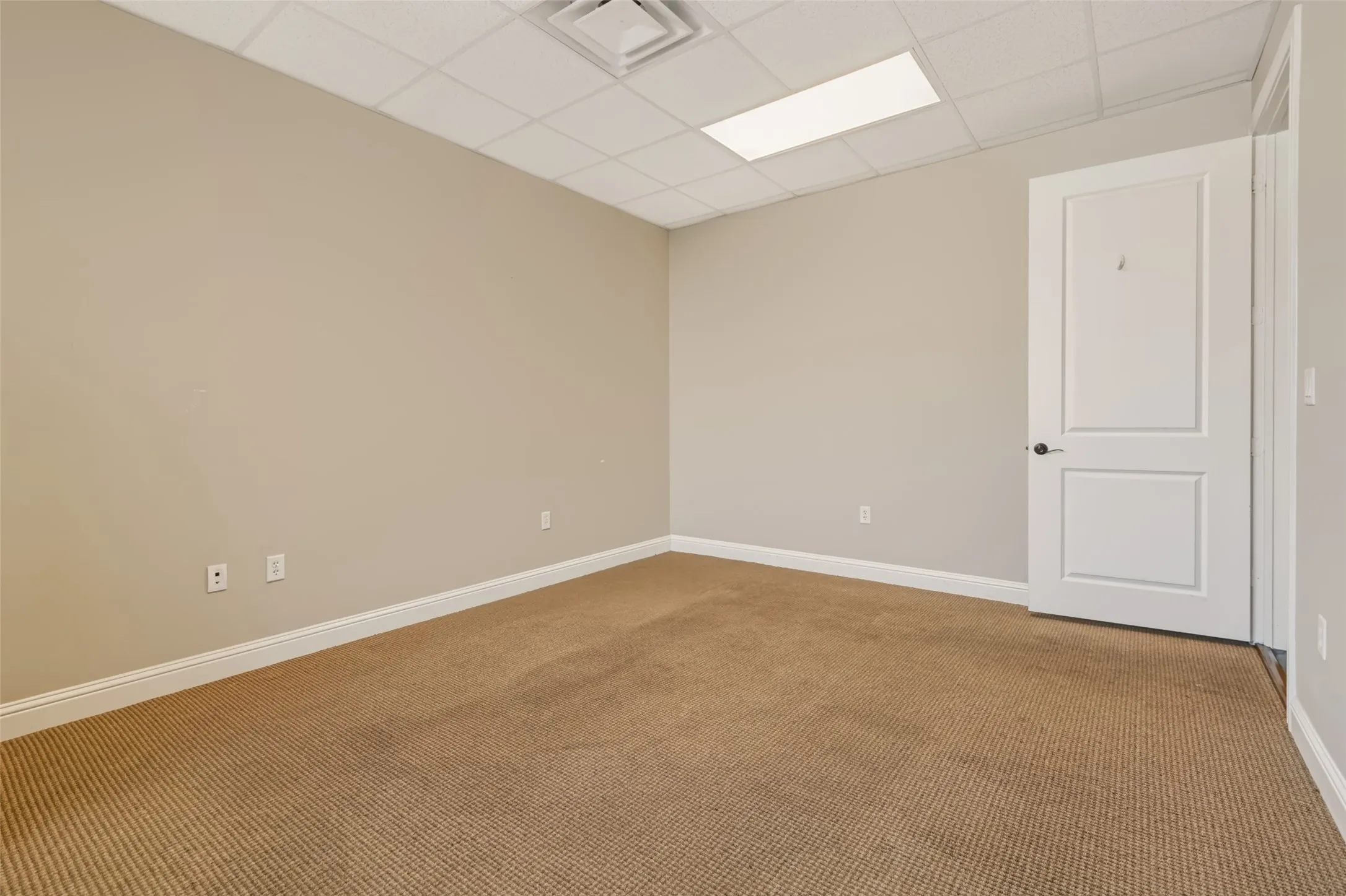 Neutral wall paint, drop ceiling with integrated lighting, textured carpet flooring, white trim, and a paneled interior door with dark hardware