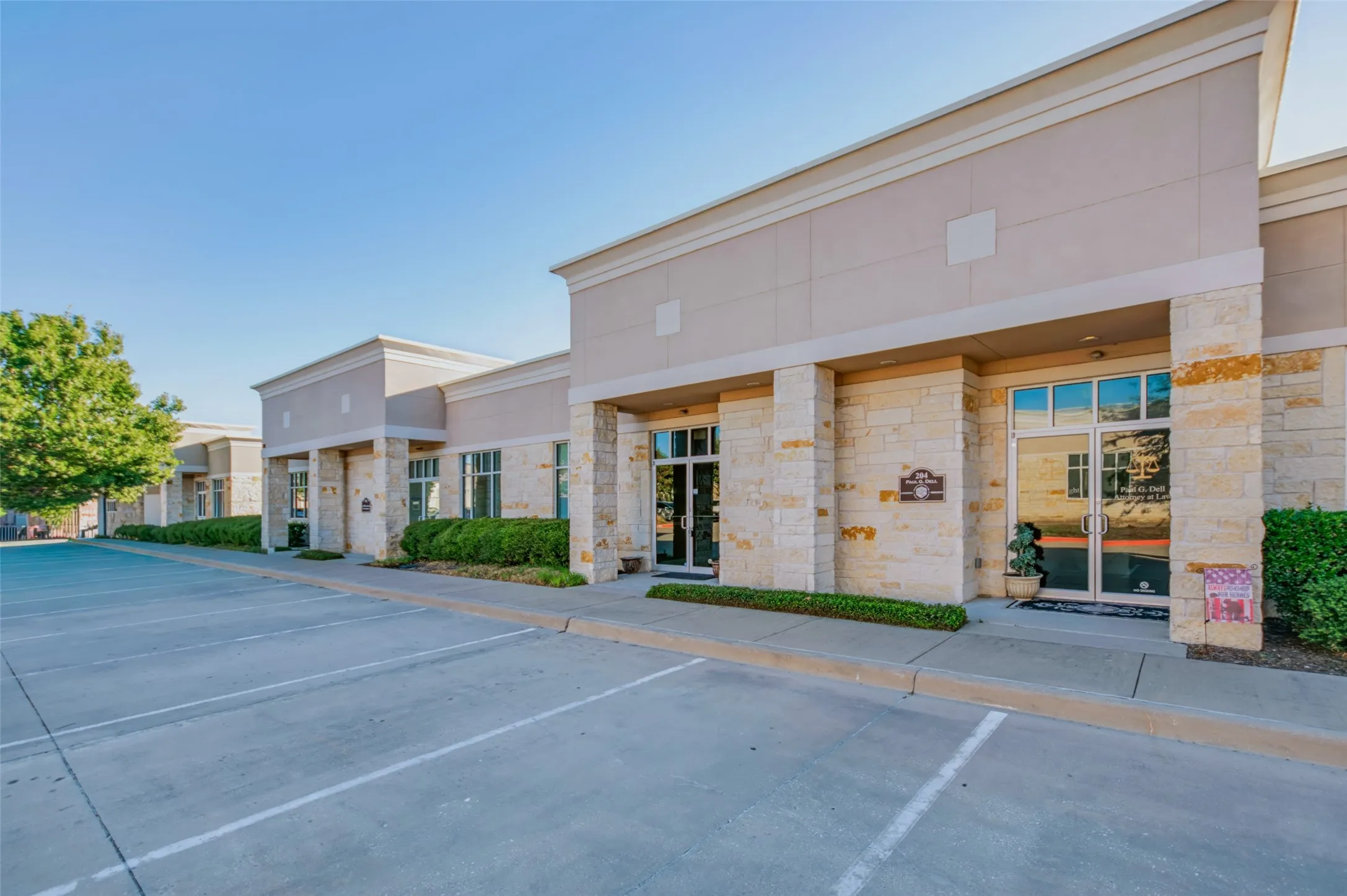 Stone and stucco facade commercial building with multiple storefronts, extensive paved parking, and mature perimeter landscaping