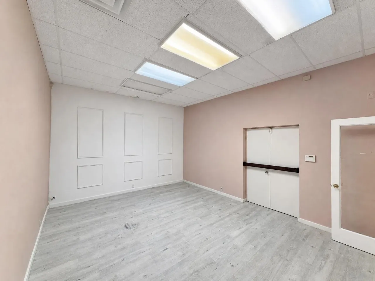 Recessed ceiling with integrated lighting, decorative wall paneling, wood-finish flooring, and a glass-paneled door