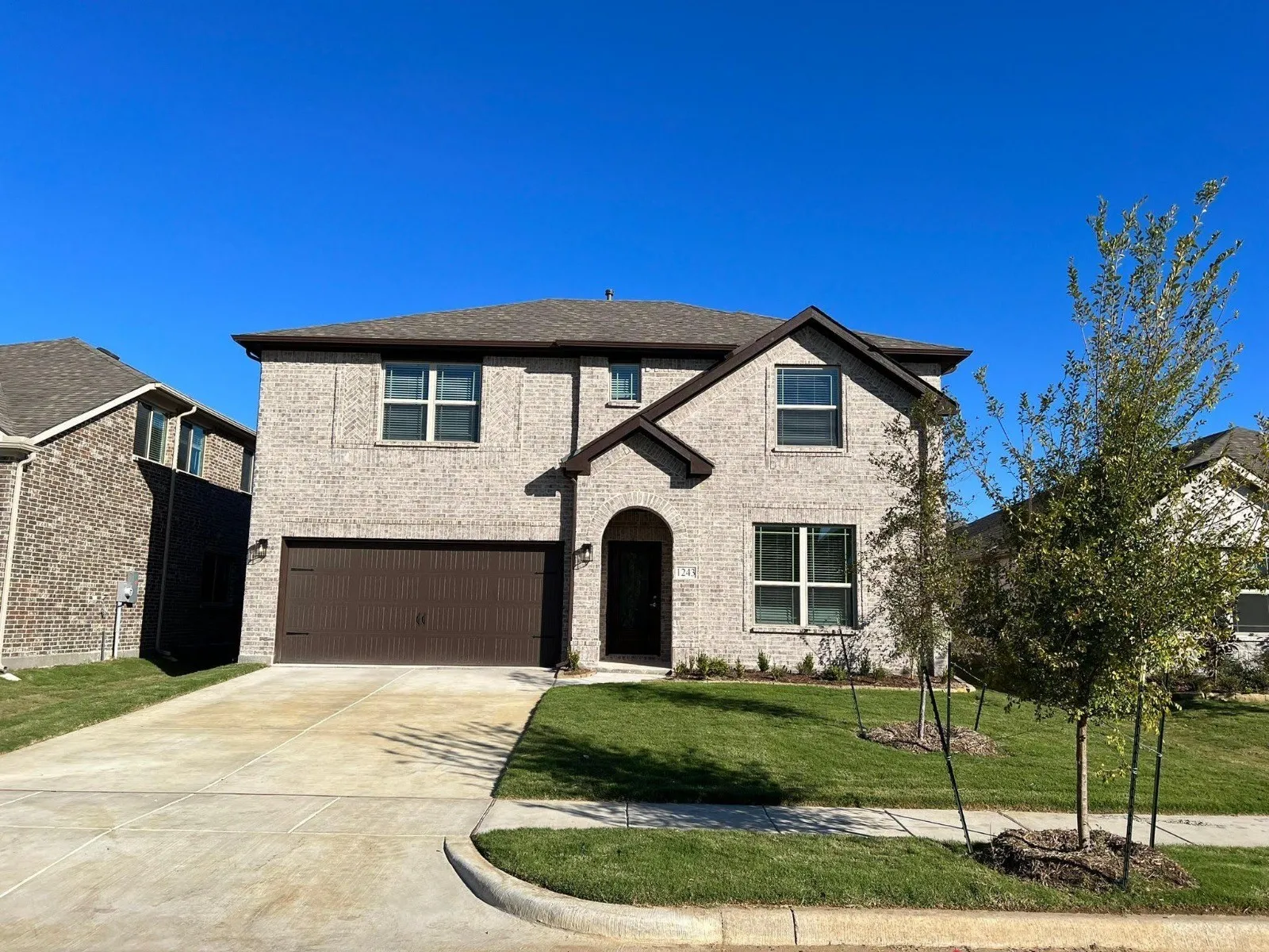 Brick facade with dark trim accents, attached two-car garage, concrete driveway, arched entry, and mature landscaping