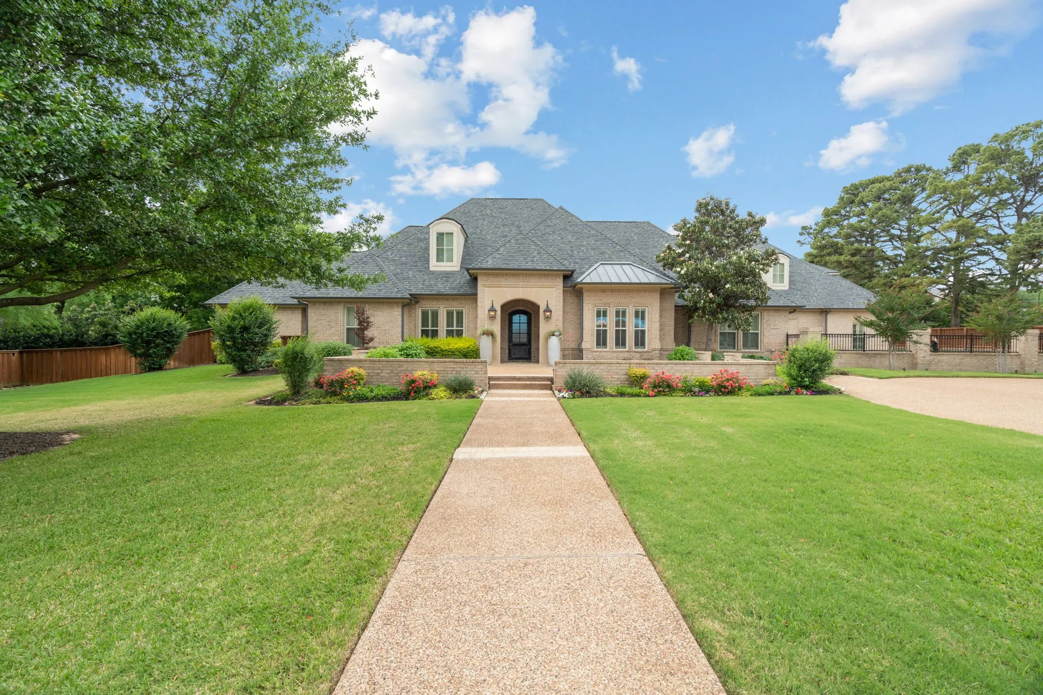 Stone façade with dormer windows, a prominent entryway, a long concrete pathway, and landscaped garden beds