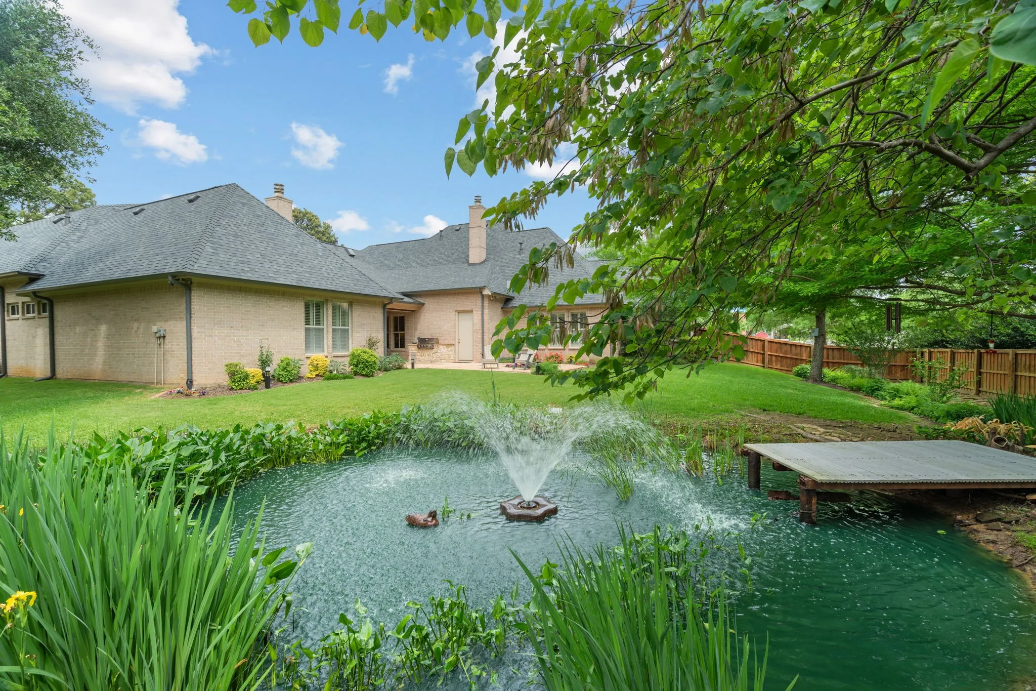 Brick exterior with dark shingle roof, expansive green lawn, mature trees, and a pond with a water feature and dock