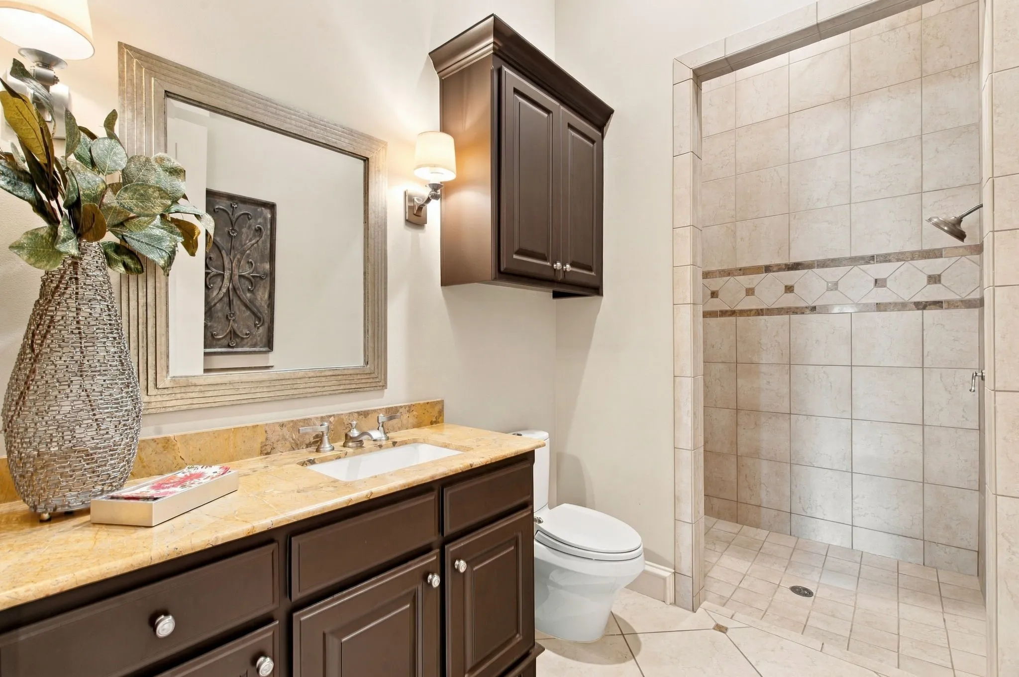 Bathroom featuring an integrated vanity sink with a light-colored countertop, dark wood-finish cabinetry, a large framed mirror, and a tiled shower with a decorative accent band