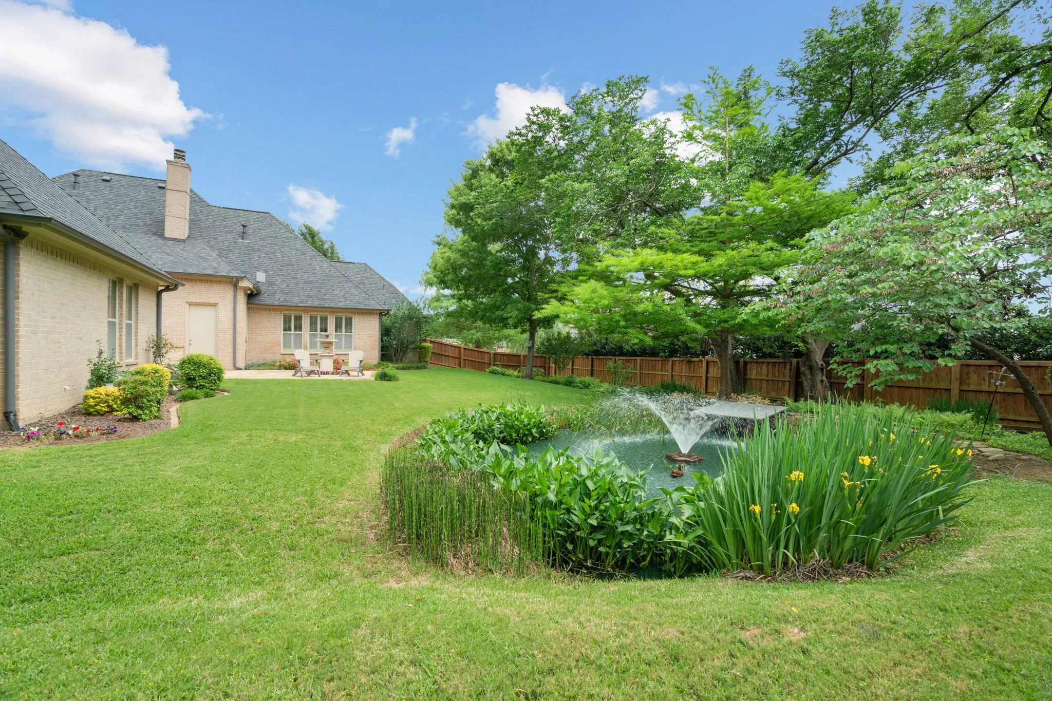 Expansive lawn with a built-in pond featuring a fountain, surrounded by mature trees, a wooden perimeter fence, and a patio area