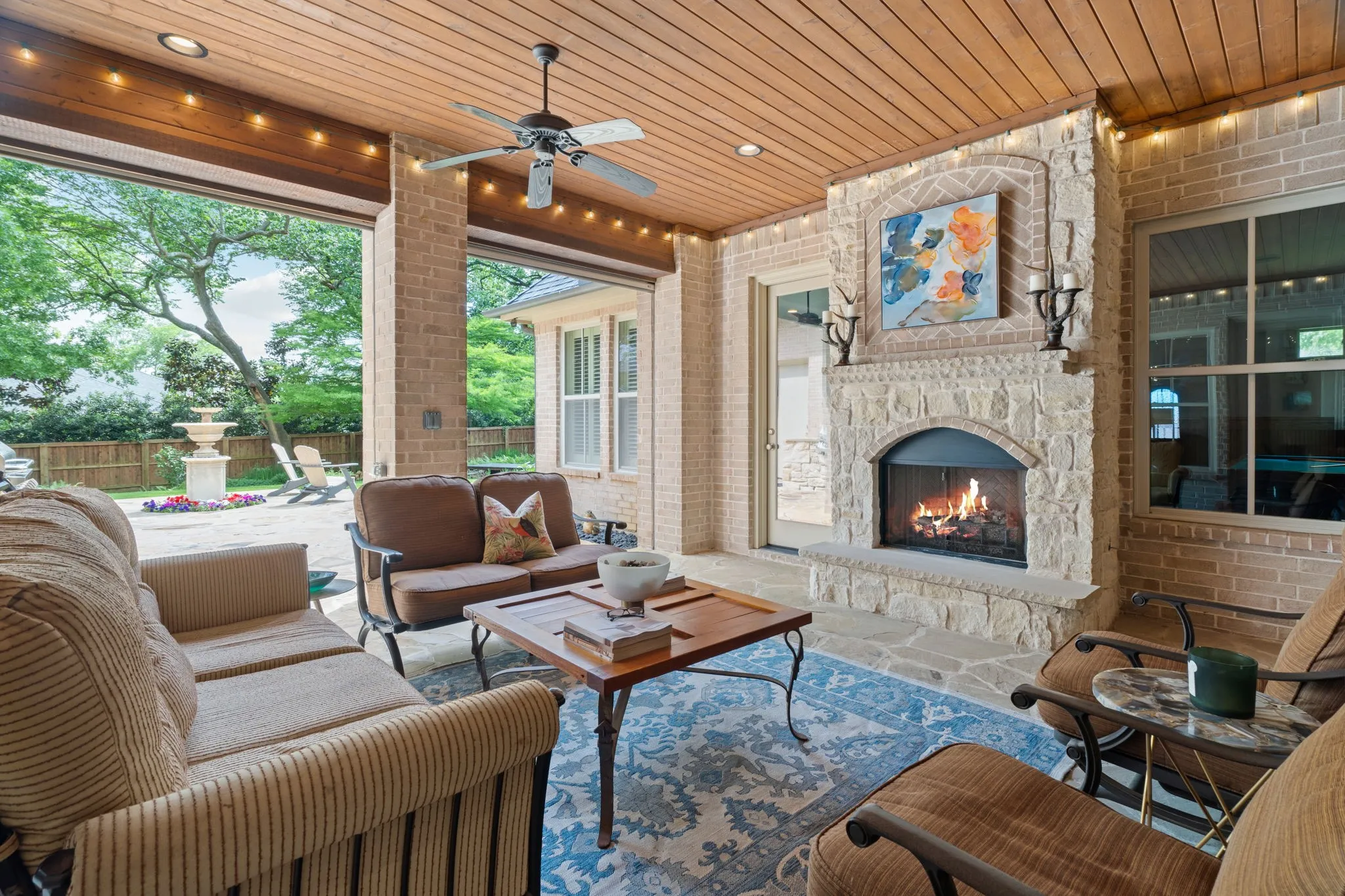 Covered patio with a wood plank ceiling and recessed lighting, featuring a stone-clad fireplace, brick columns, and a ceiling fan
