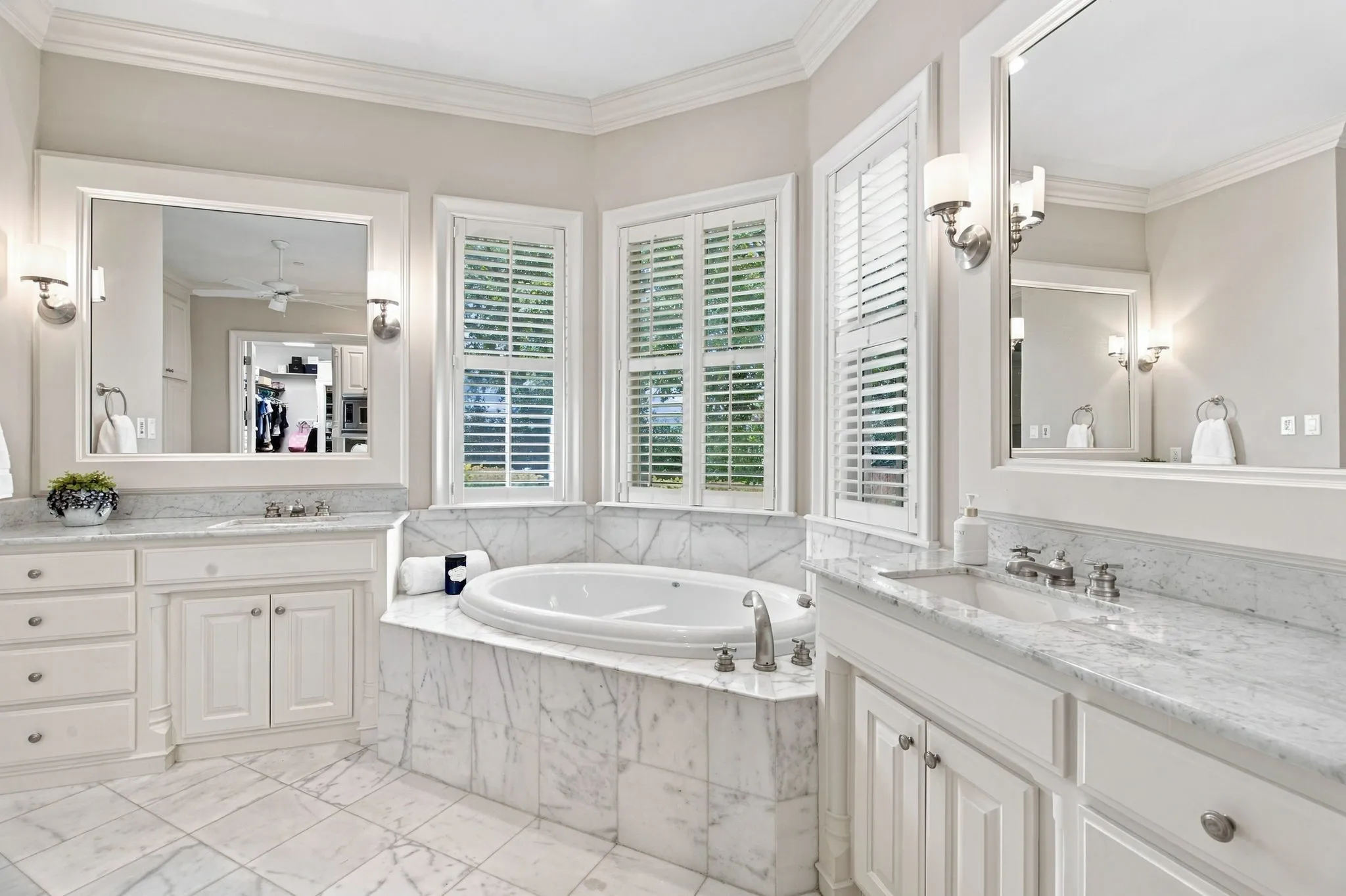 Dual vanity with integrated sinks, white cabinetry, a built-in soaking tub, marble-patterned tile flooring, and shuttered windows