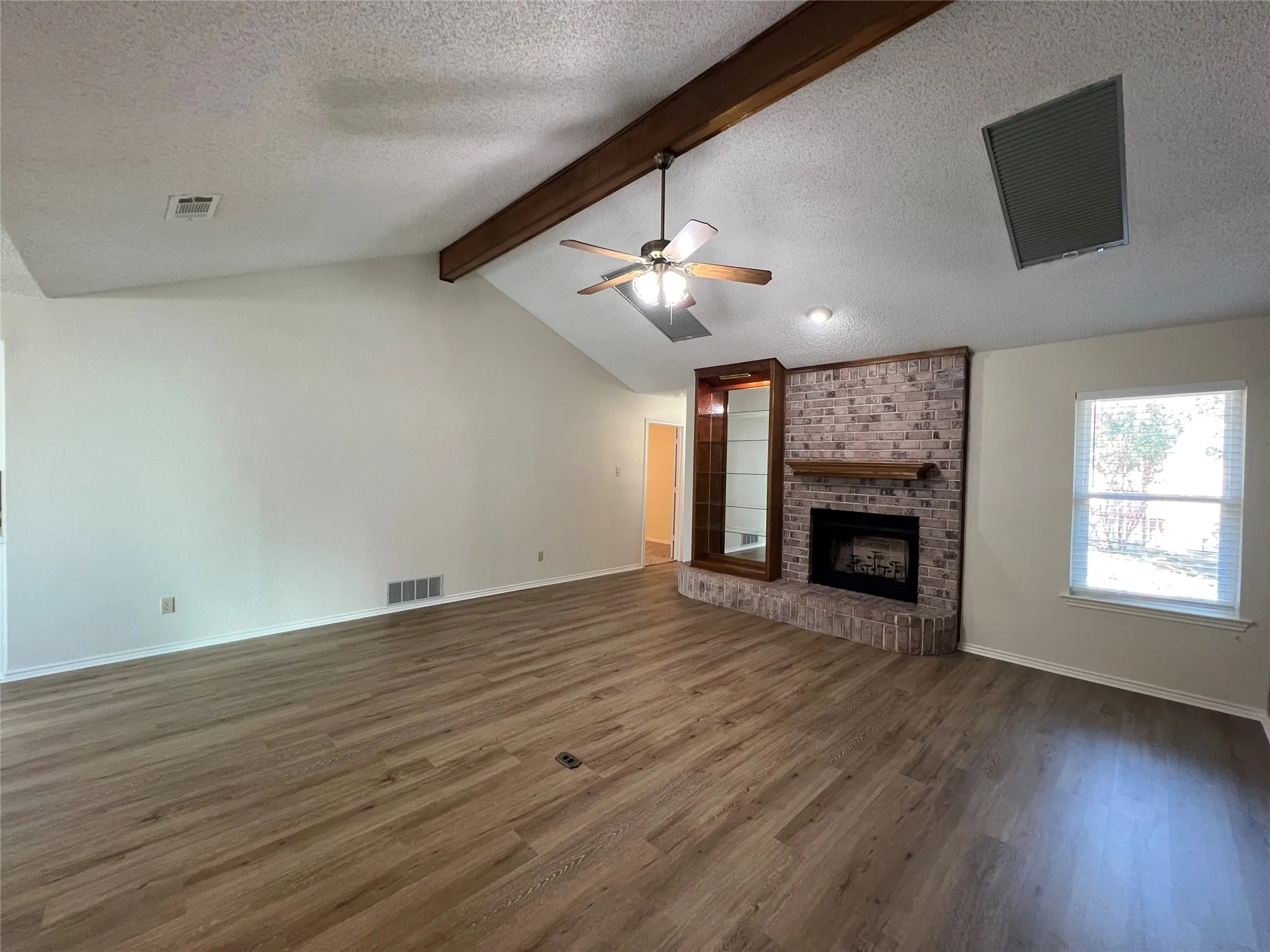 Vaulted ceiling with exposed wood beam, wood-finish flooring, brick-surround fireplace with wood mantel, built-in display cabinetry, and ceiling fan