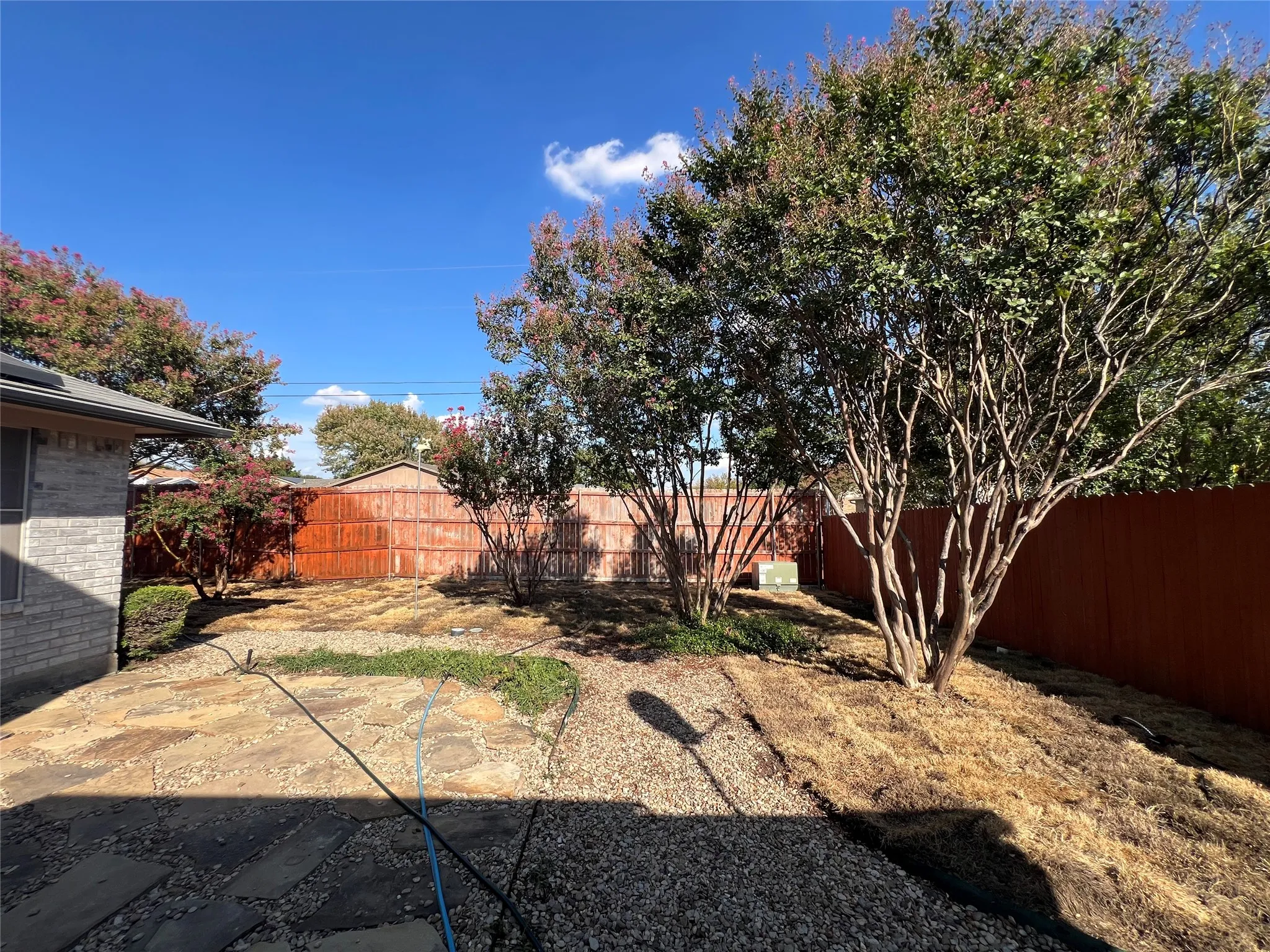 Expansive outdoor space featuring gravel and stone paver pathways, mature trees with red foliage, and a wood privacy fence