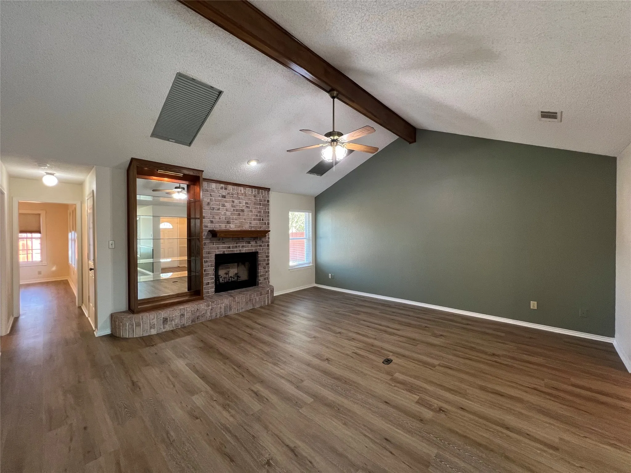 Vaulted ceiling with wood beam accents, brick fireplace with wood mantel, wood-finish flooring, built-in shelving, and a ceiling fan