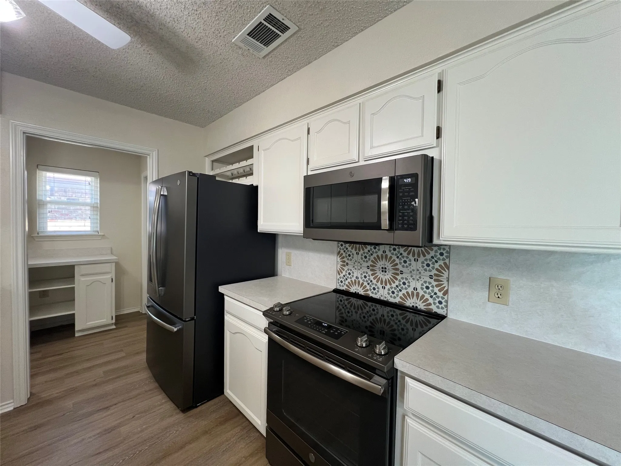 White cabinetry, wood-finish flooring, stainless steel microwave, black electric range, and patterned tile backsplash
