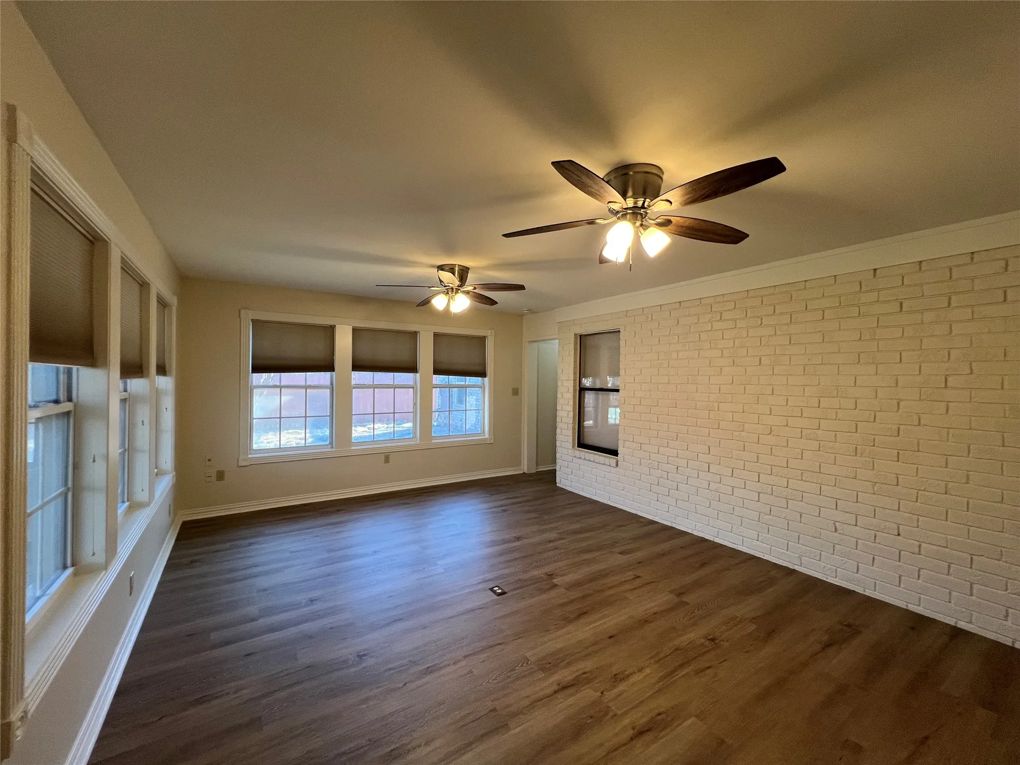 Wood-finish flooring, white painted brick accent wall, multiple windows with blinds, and two ceiling fans with integrated lighting